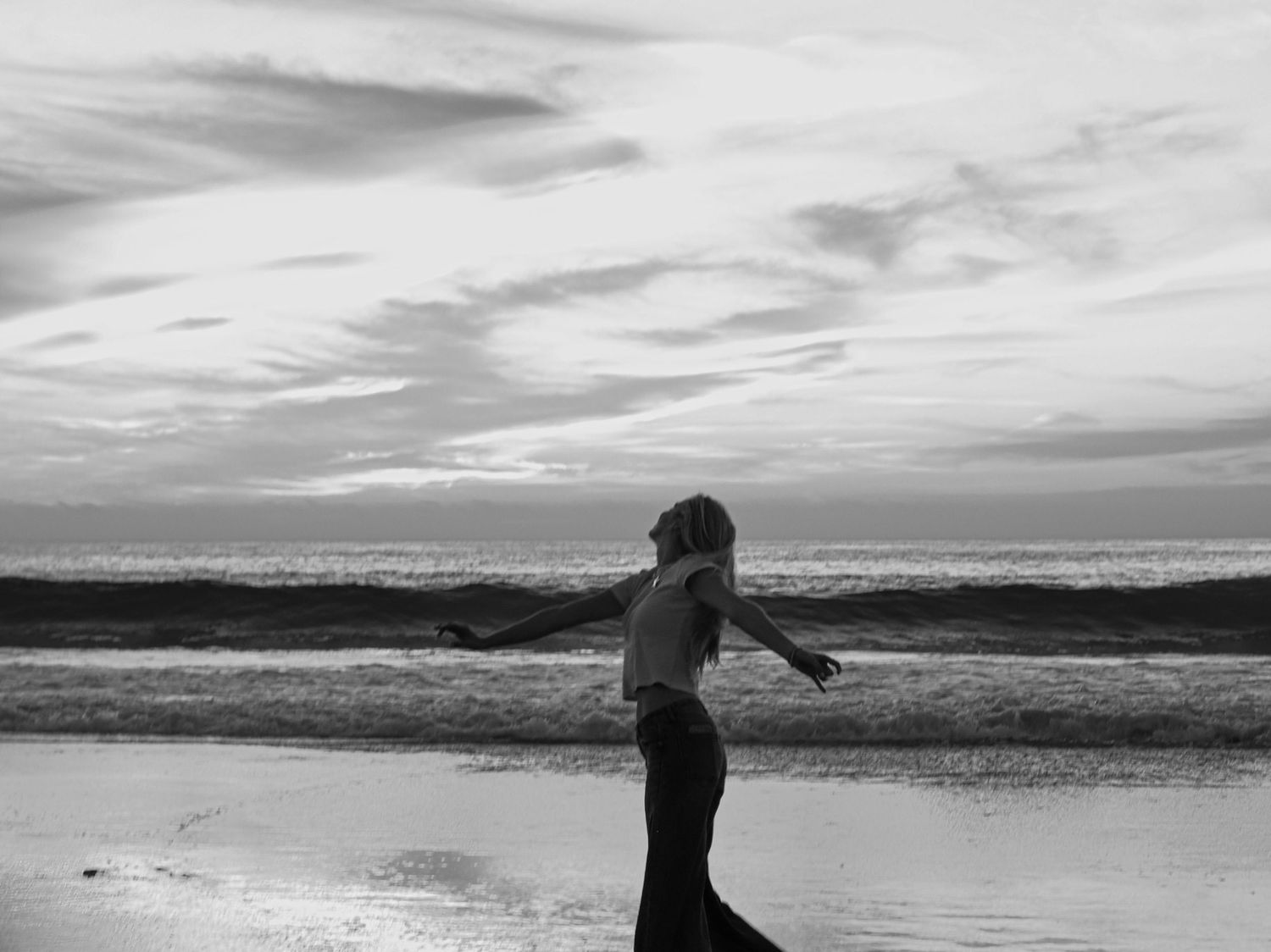A silhouetted figure spins on a beach at dusk with dramatic clouds overhead in black and white.