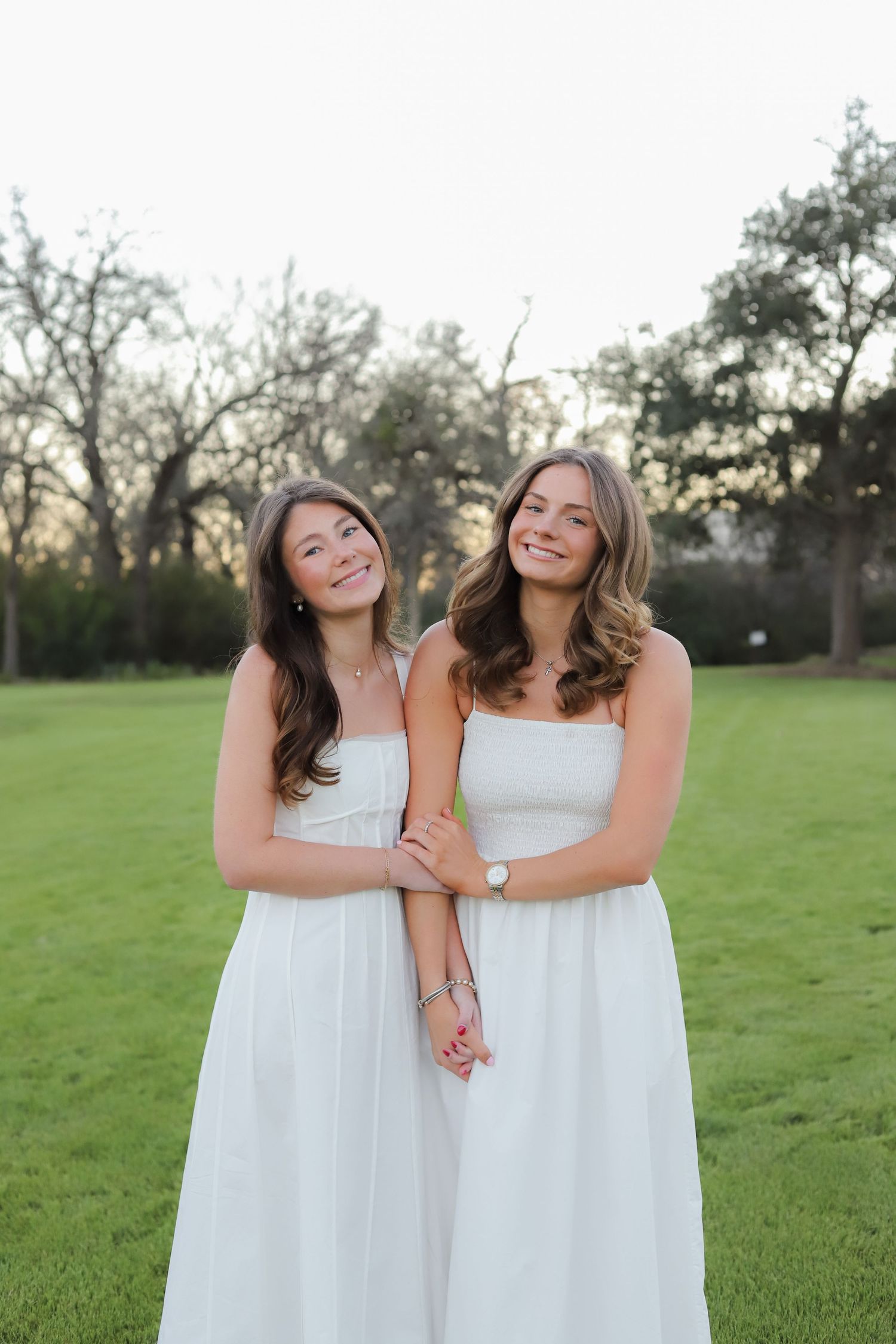 Two friends in white dresses embrace while laughing on a green lawn during golden hour.