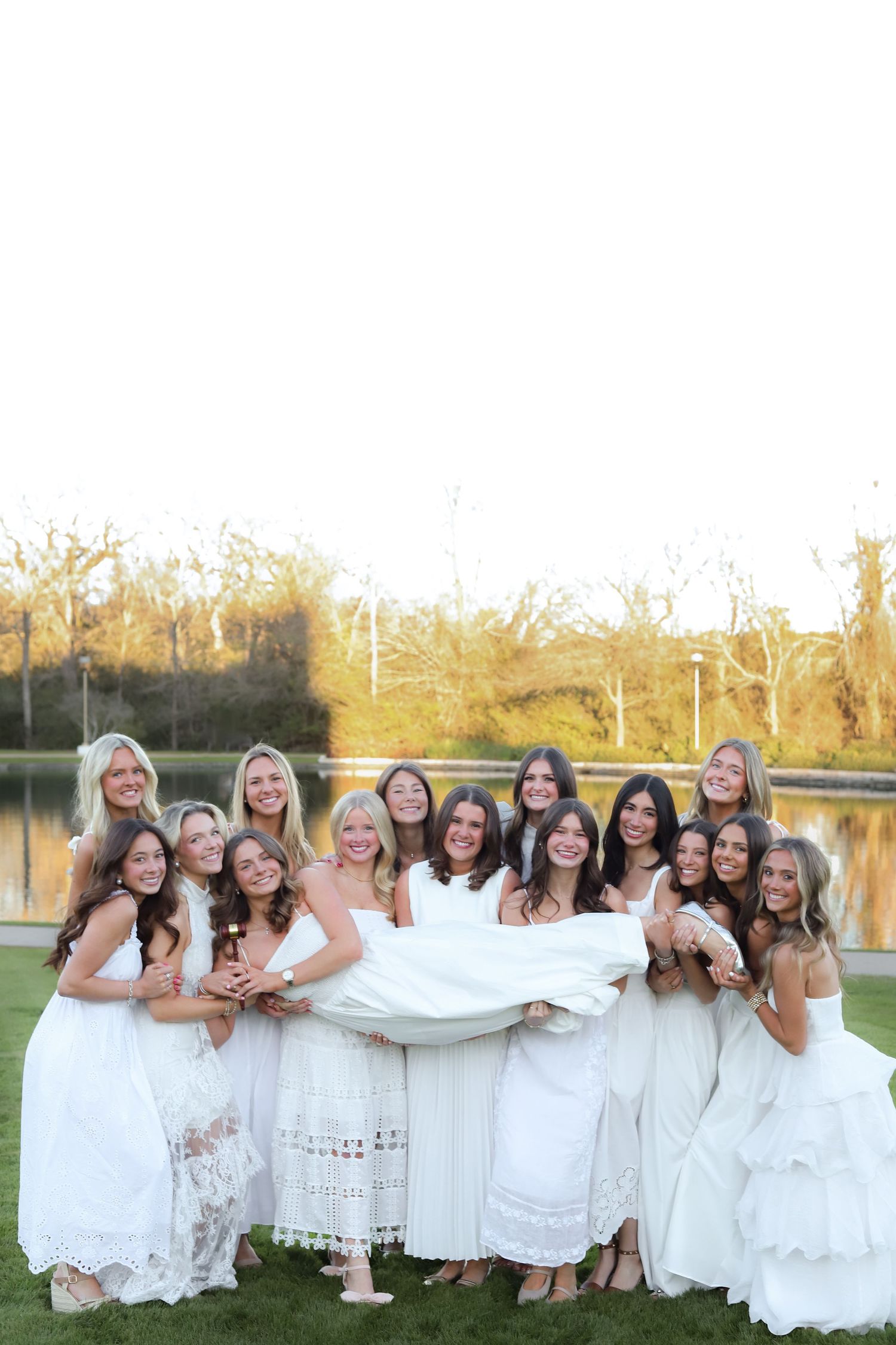 A large group in coordinated white dresses poses joyfully together on grass during golden hour.