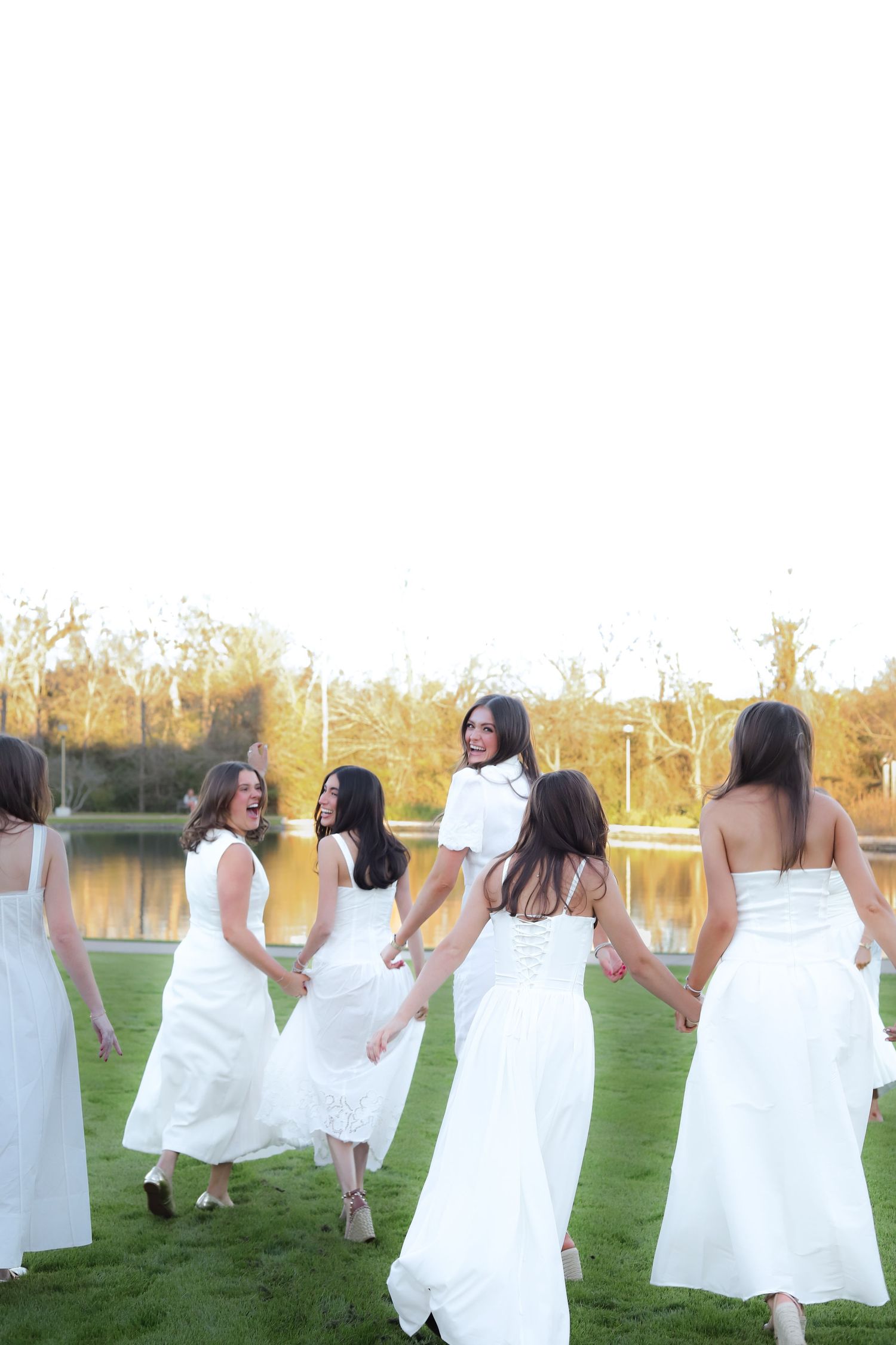 Friends in white dresses dance and twirl together in a grassy field at sunset.