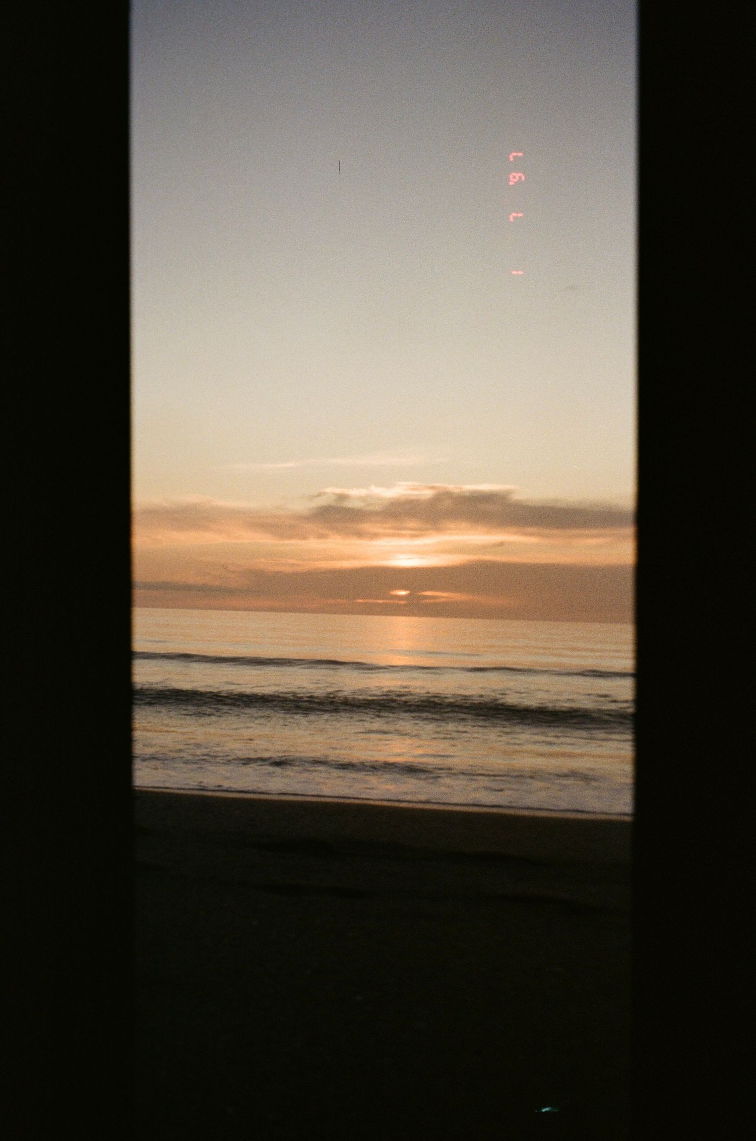 Peaceful ocean sunset view through a dark window frame with gentle waves rolling onto shore.
