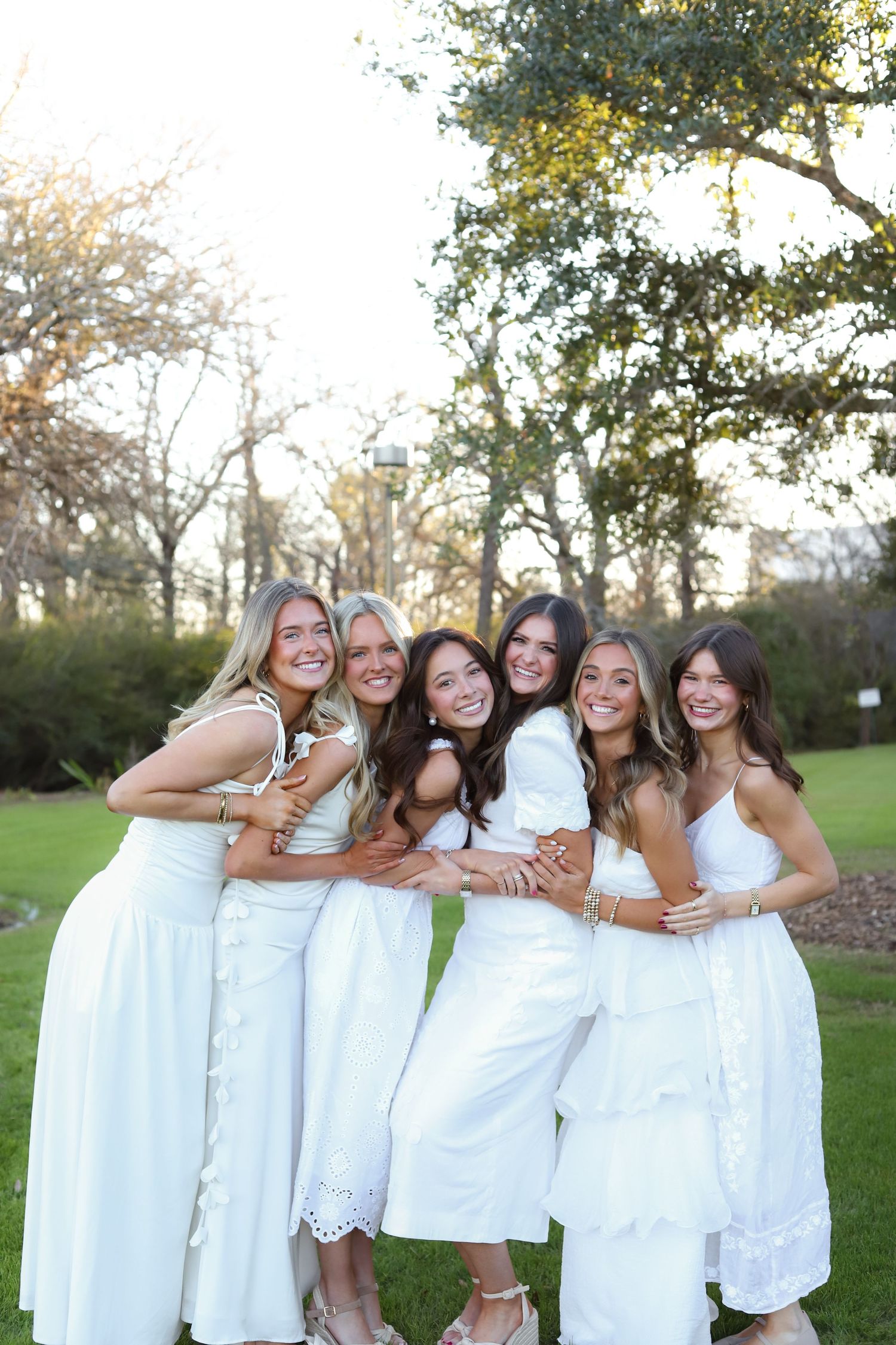 A group in matching white dresses pose together outdoors in a playful line formation.