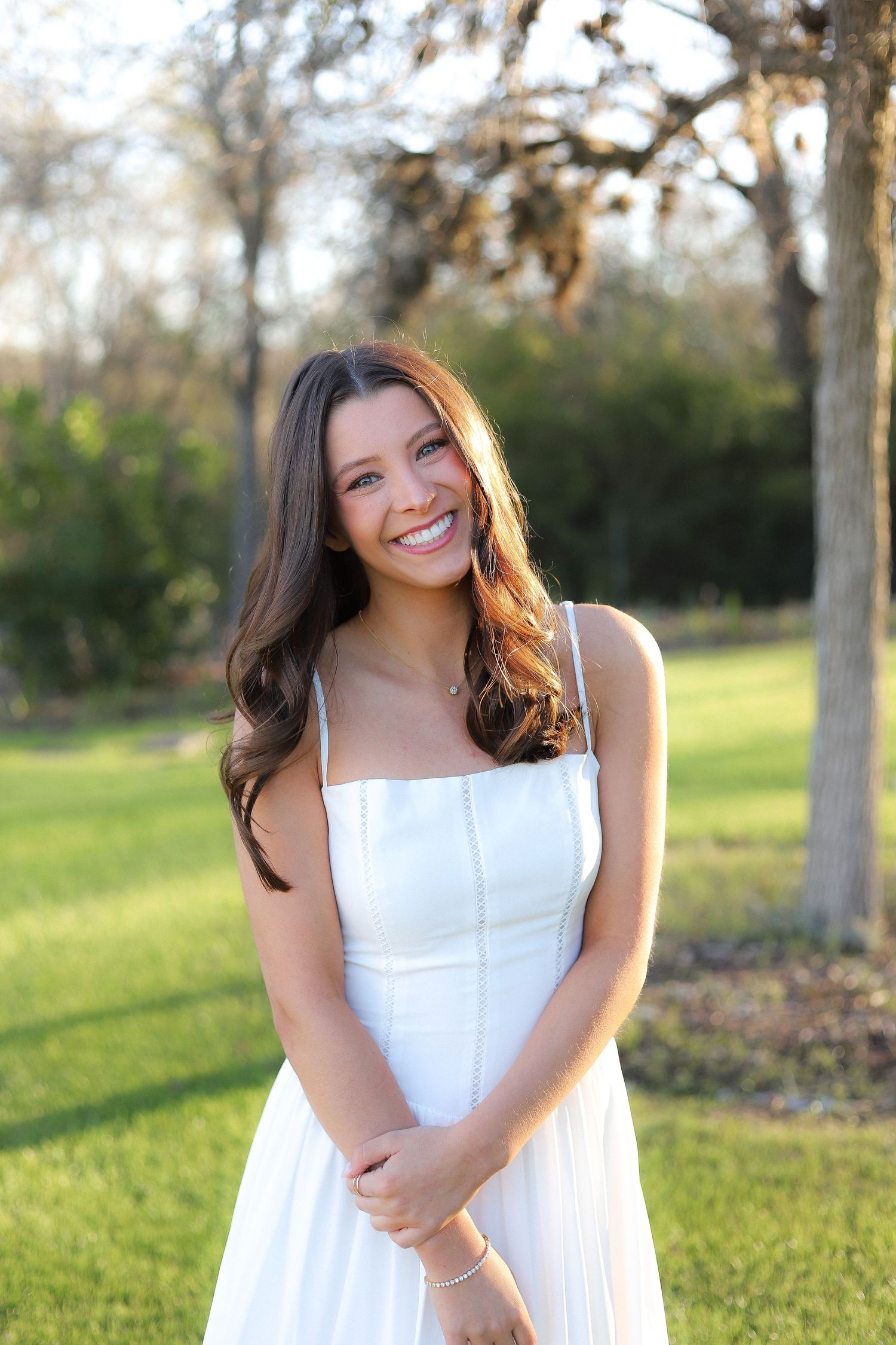 A person in a white sundress poses joyfully in a grassy outdoor setting during golden hour.