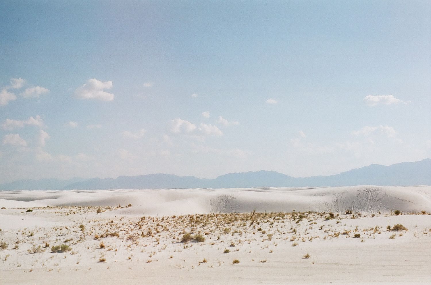 Rolling white sand dunes stretch toward distant mountains under a pale blue sky.