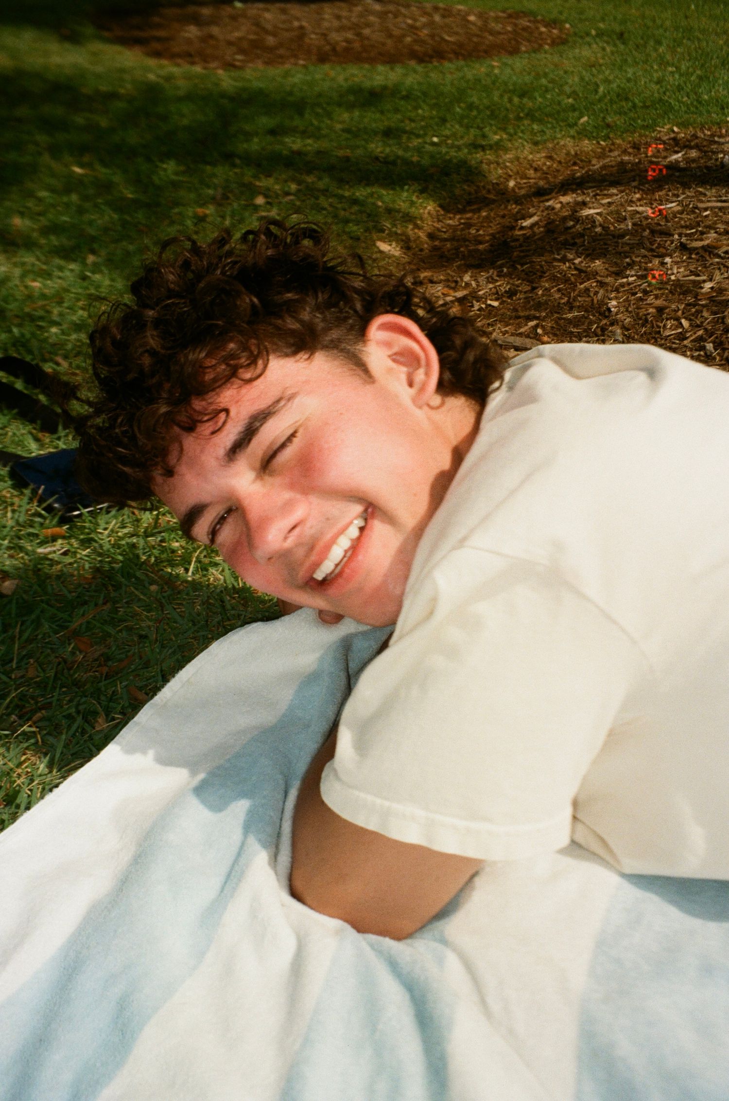 A person lying on white fabric outdoors smiles while resting on grass in natural lighting.