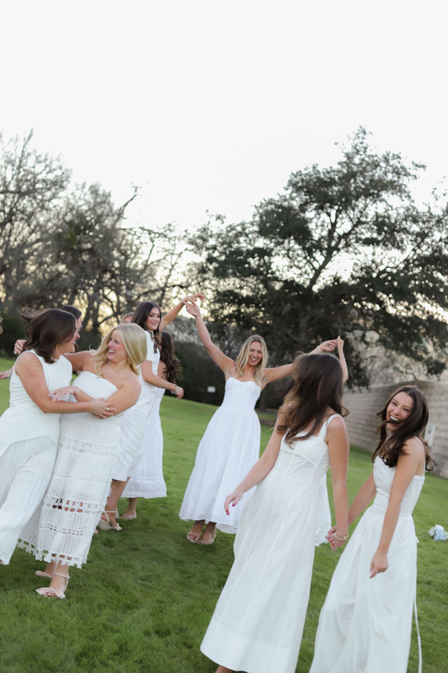 Friends in coordinating white dresses dance and celebrate together on a grassy field at sunset.