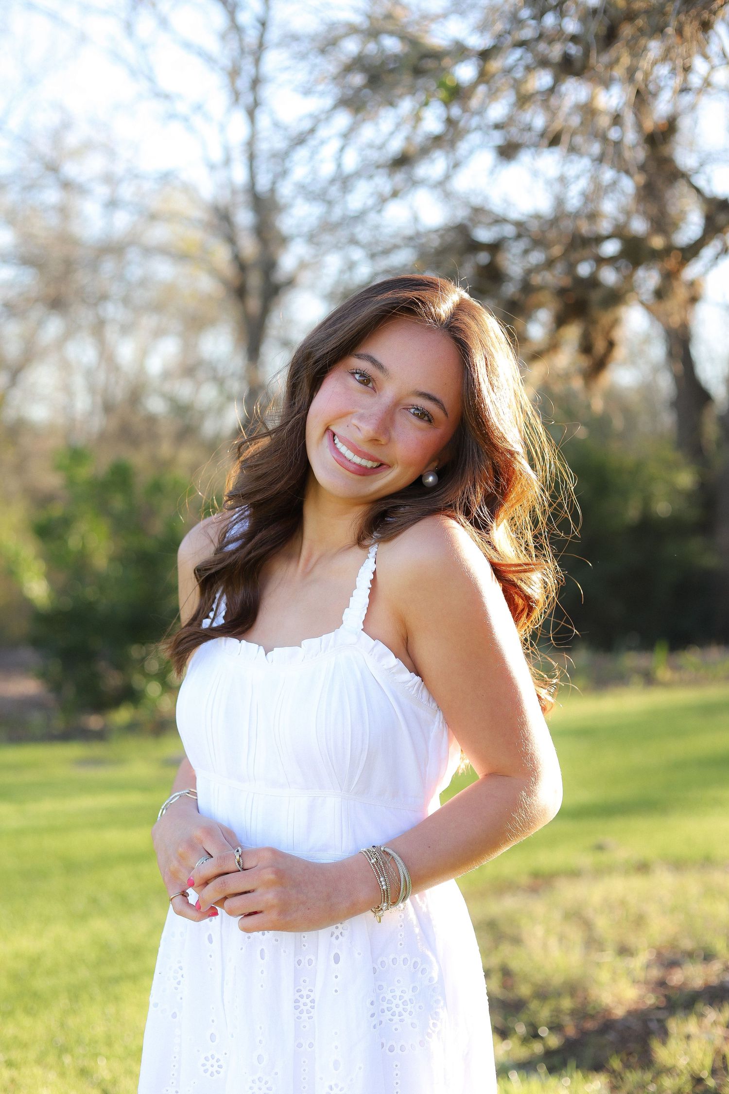 A person in a white sundress poses gracefully against a natural outdoor backdrop with sunlit trees.