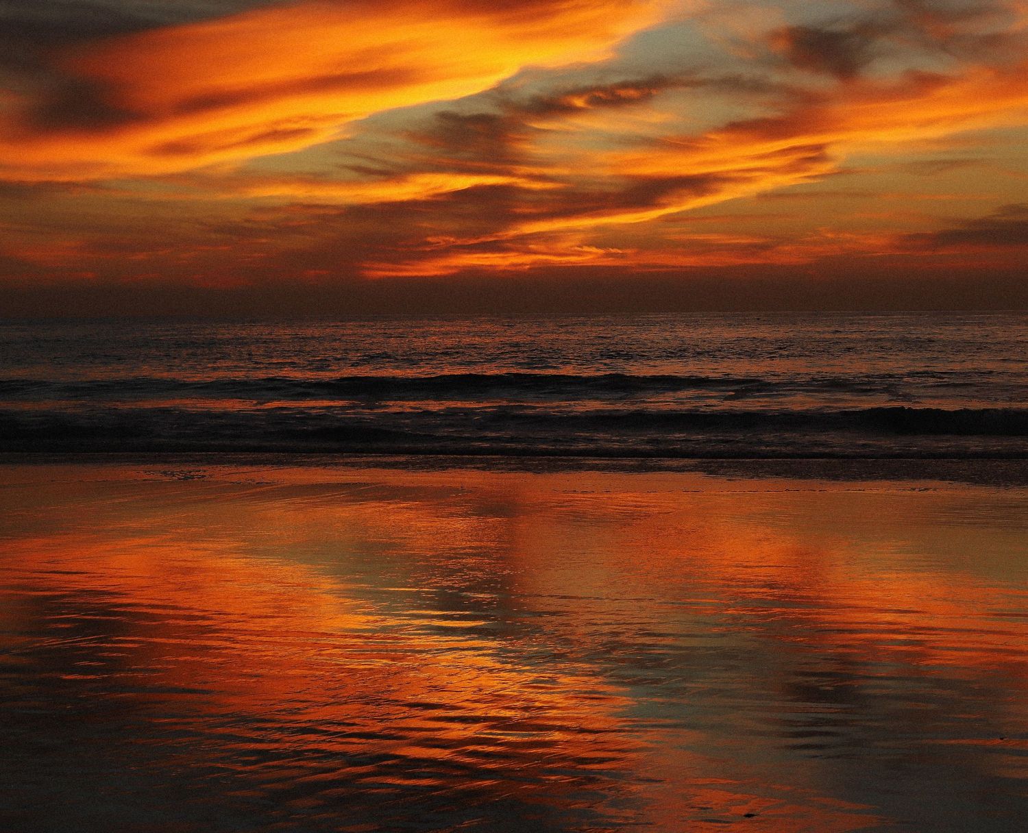 Dramatic orange sunset reflects on wet beach sand as waves roll in under fiery clouds over the ocean.