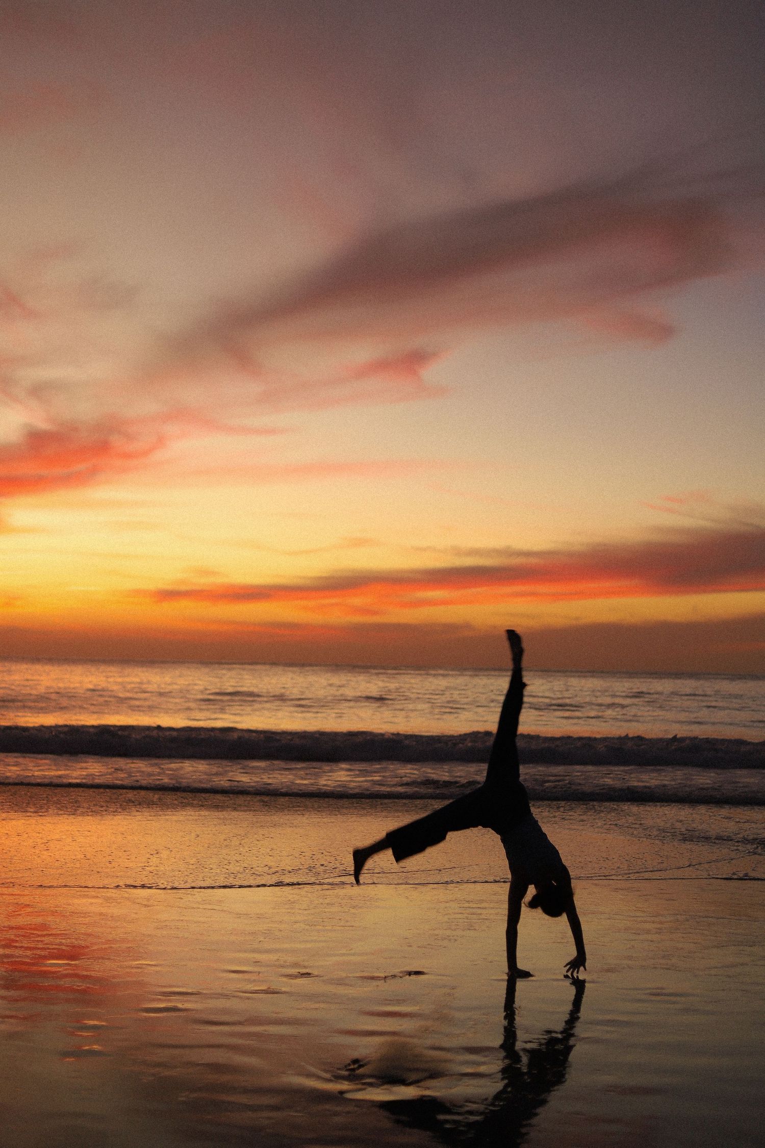 Silhouetted figures perform acrobatic poses on a beach at sunset with vibrant orange and pink sky reflecting on the water.