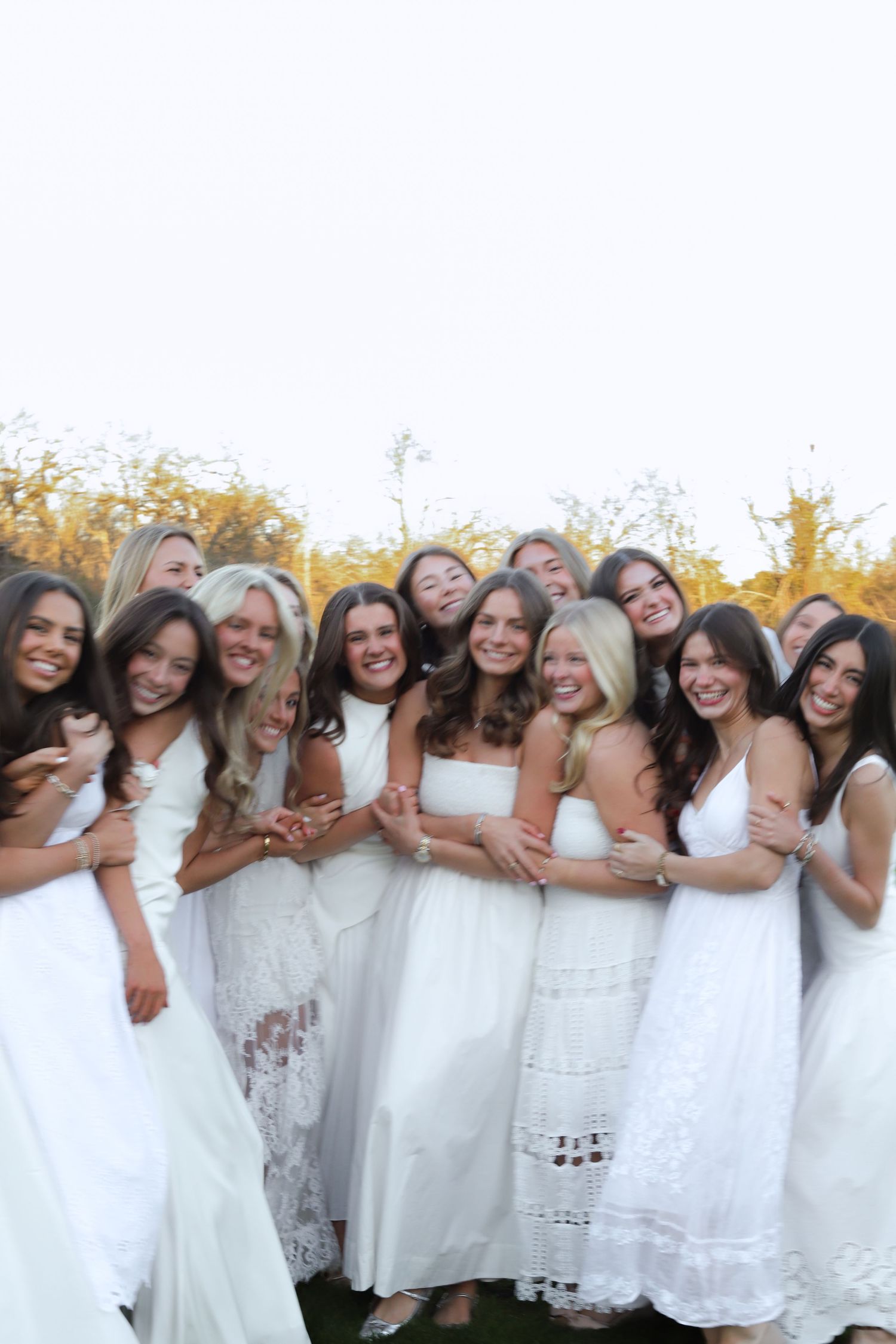 A group wearing white dresses lean together in a row for a cheerful outdoor photo.