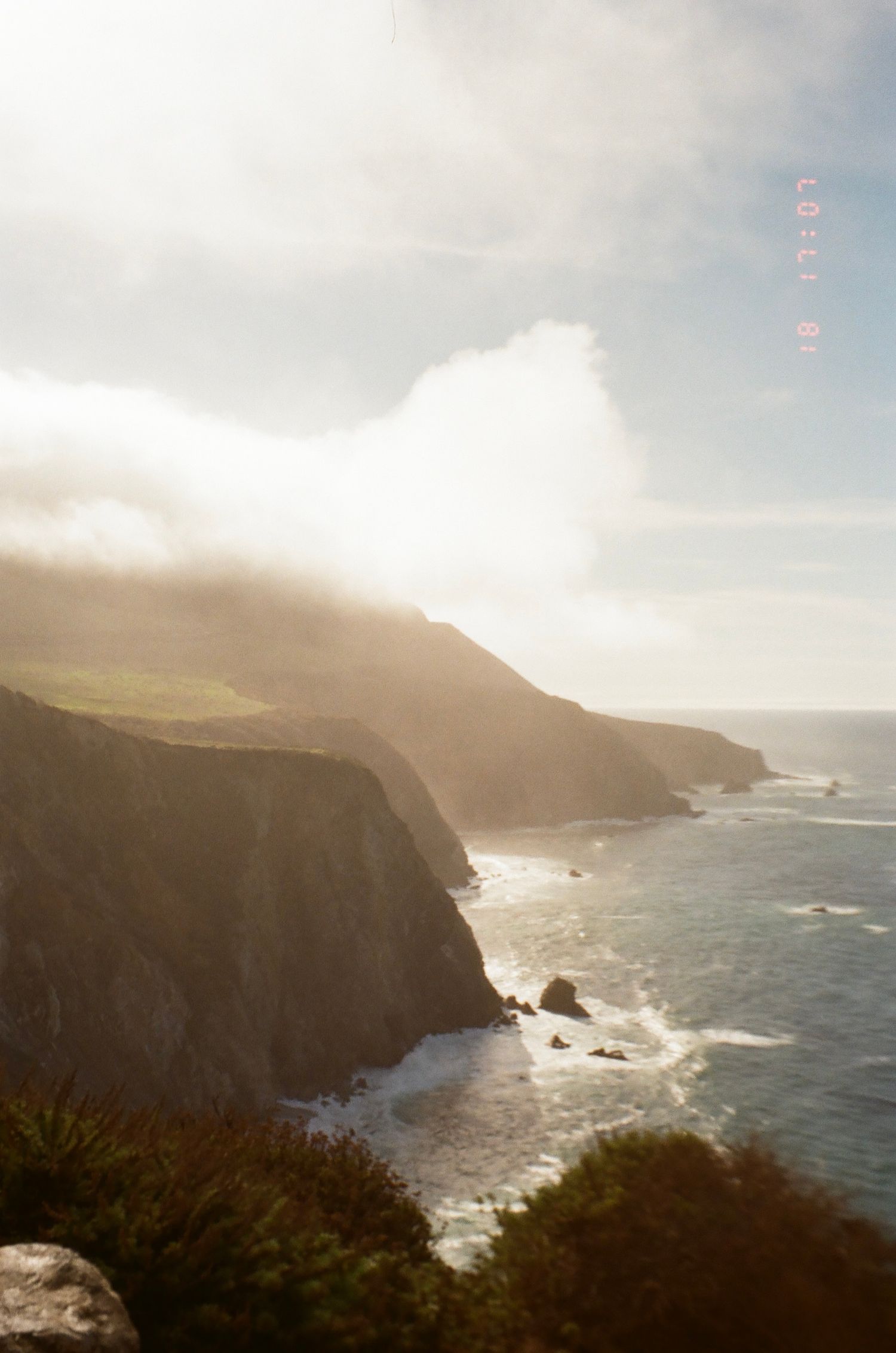 Dramatic coastal cliffs with crashing waves and misty mountains along a rugged shoreline at sunset.