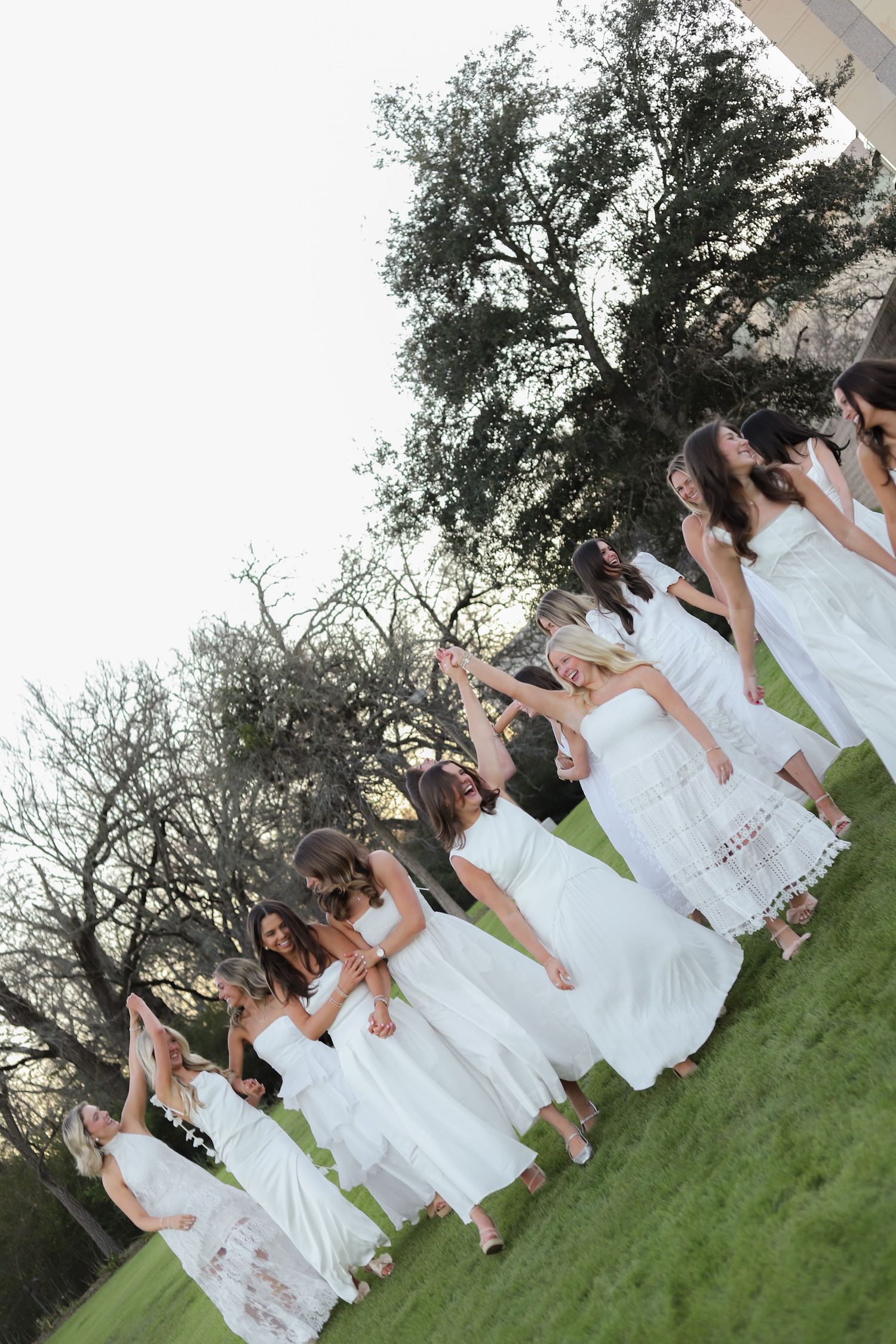 A large group poses together on grass wearing coordinated white dresses for an outdoor gathering.