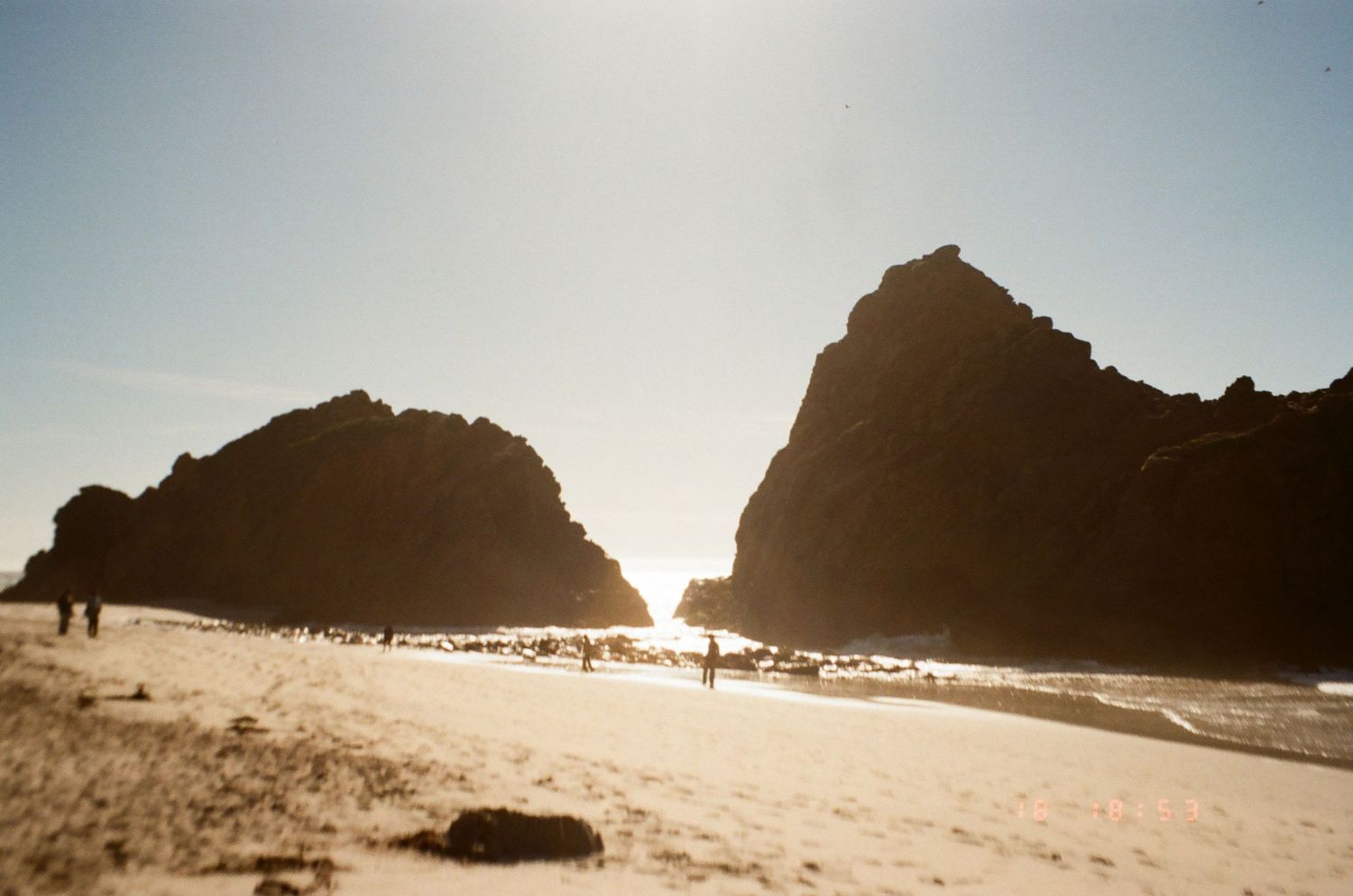 Towering rock formations create a natural gateway on a sandy beach as sunlight streams between them.