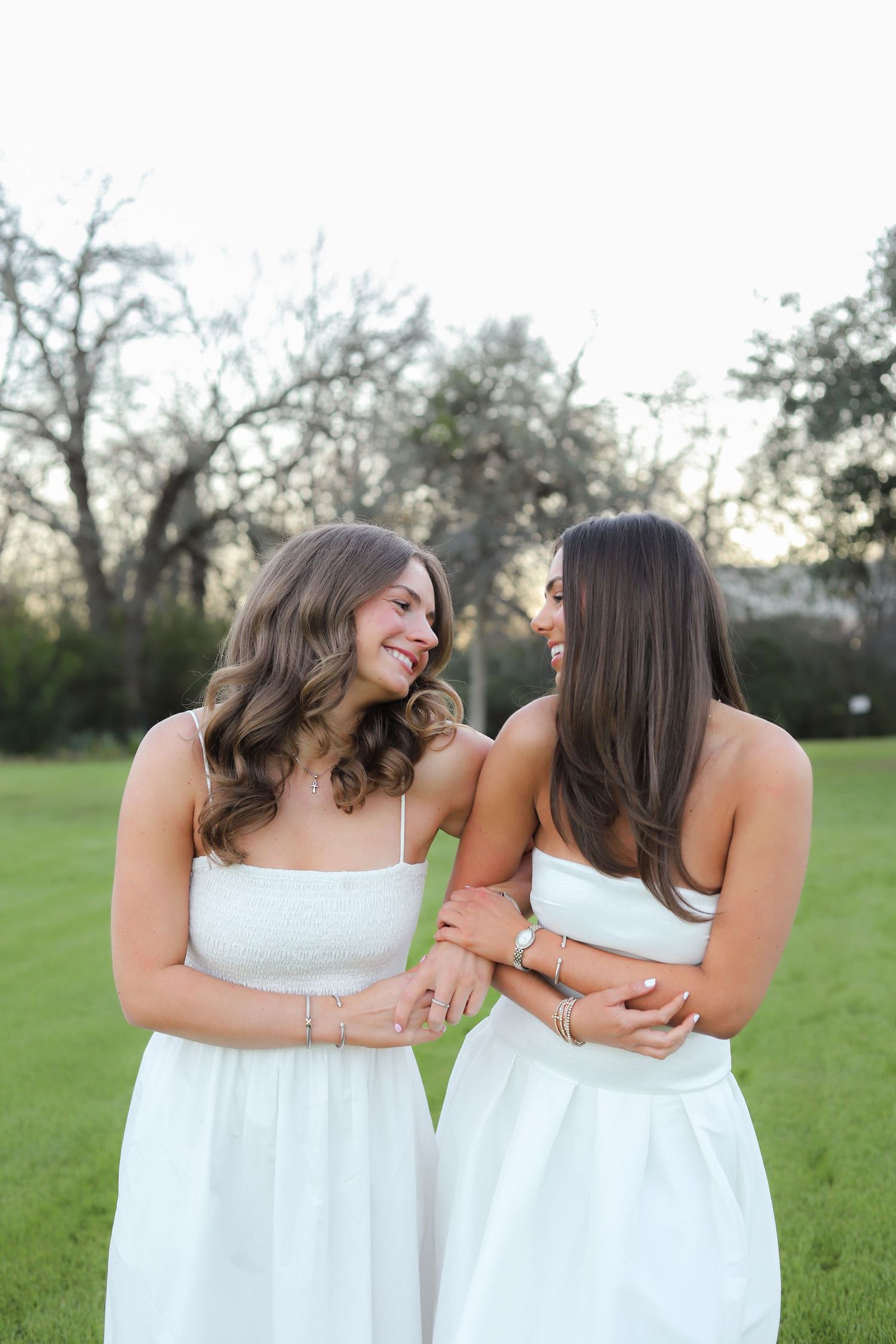 Two women in white dresses share a joyful moment together on a grassy field at sunset.