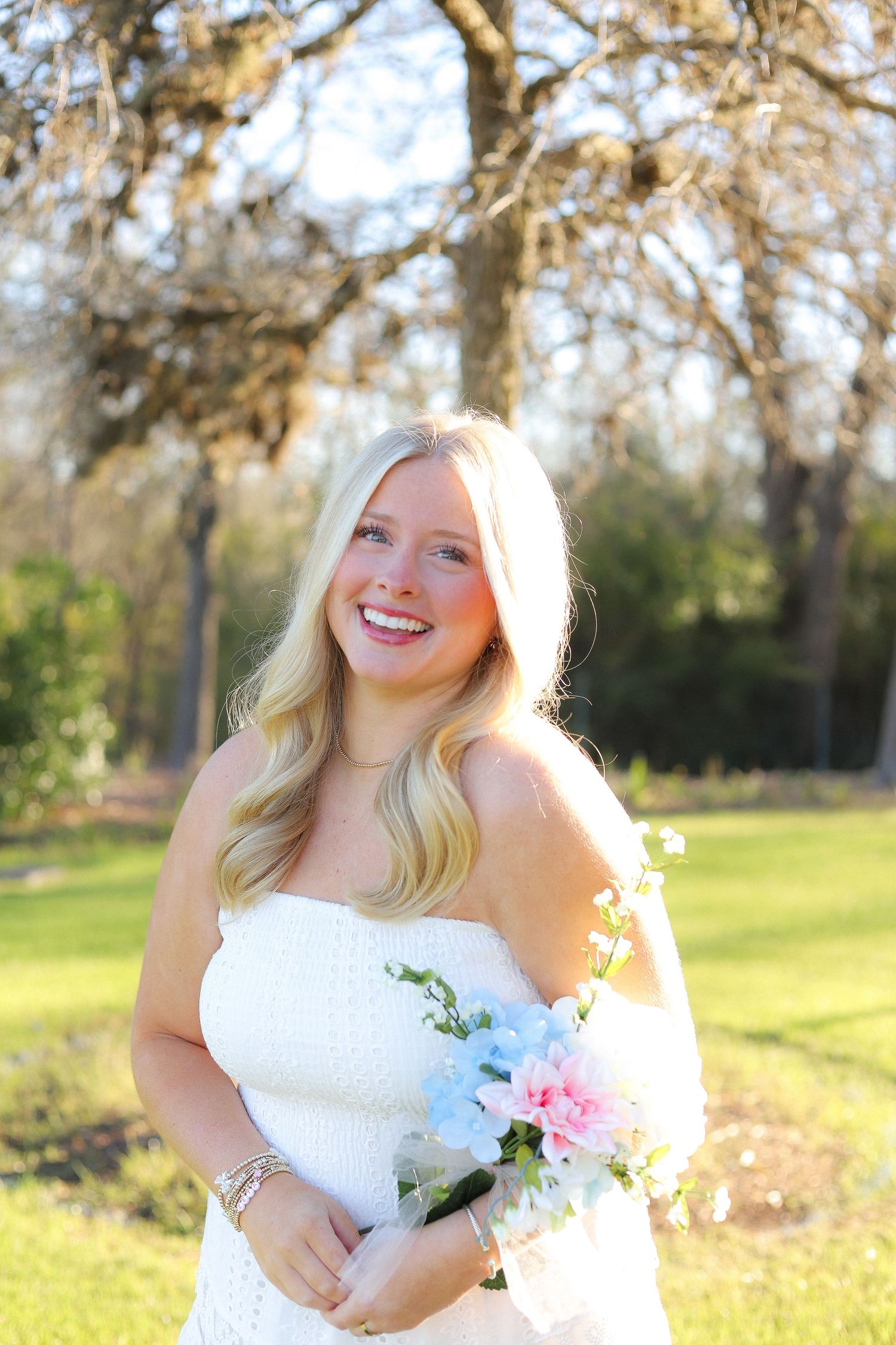 A person in a white dress holding a bouquet poses in a sunny outdoor garden setting with blooming trees.