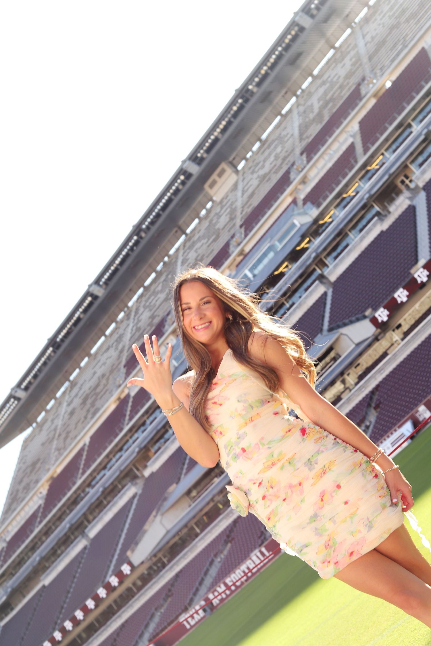 A woman in a light-colored dress waves while standing on a football field.