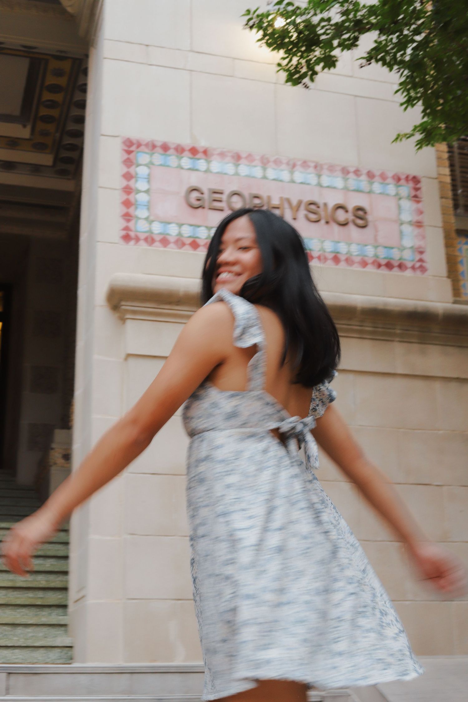 A person wearing a light gray dress spins playfully in front of a building with decorative architecture.