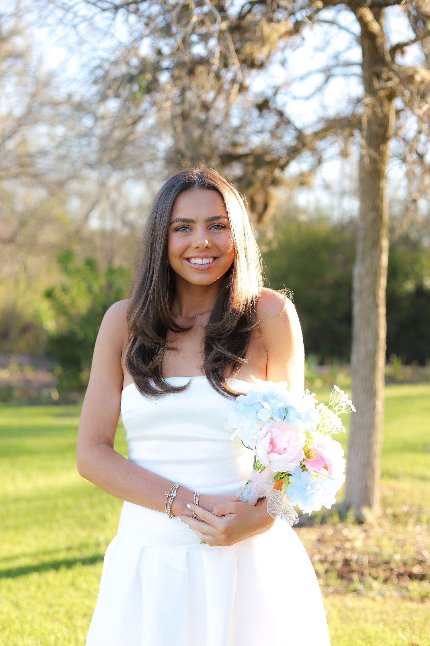 Woman in white dress holds pink flowers in outdoor garden setting at sunset.