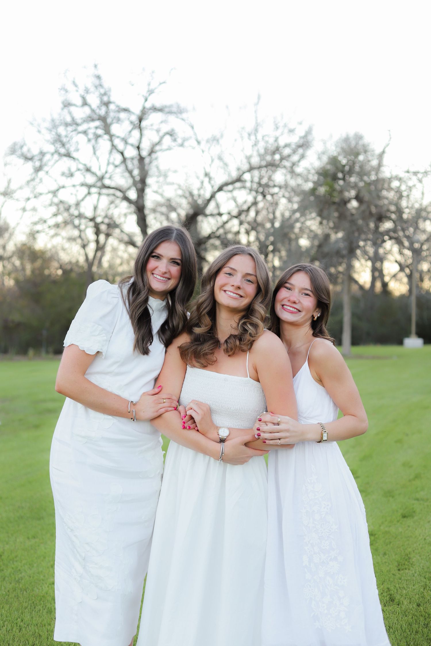 Three people in white dresses embrace and laugh together on a grassy field during golden hour.