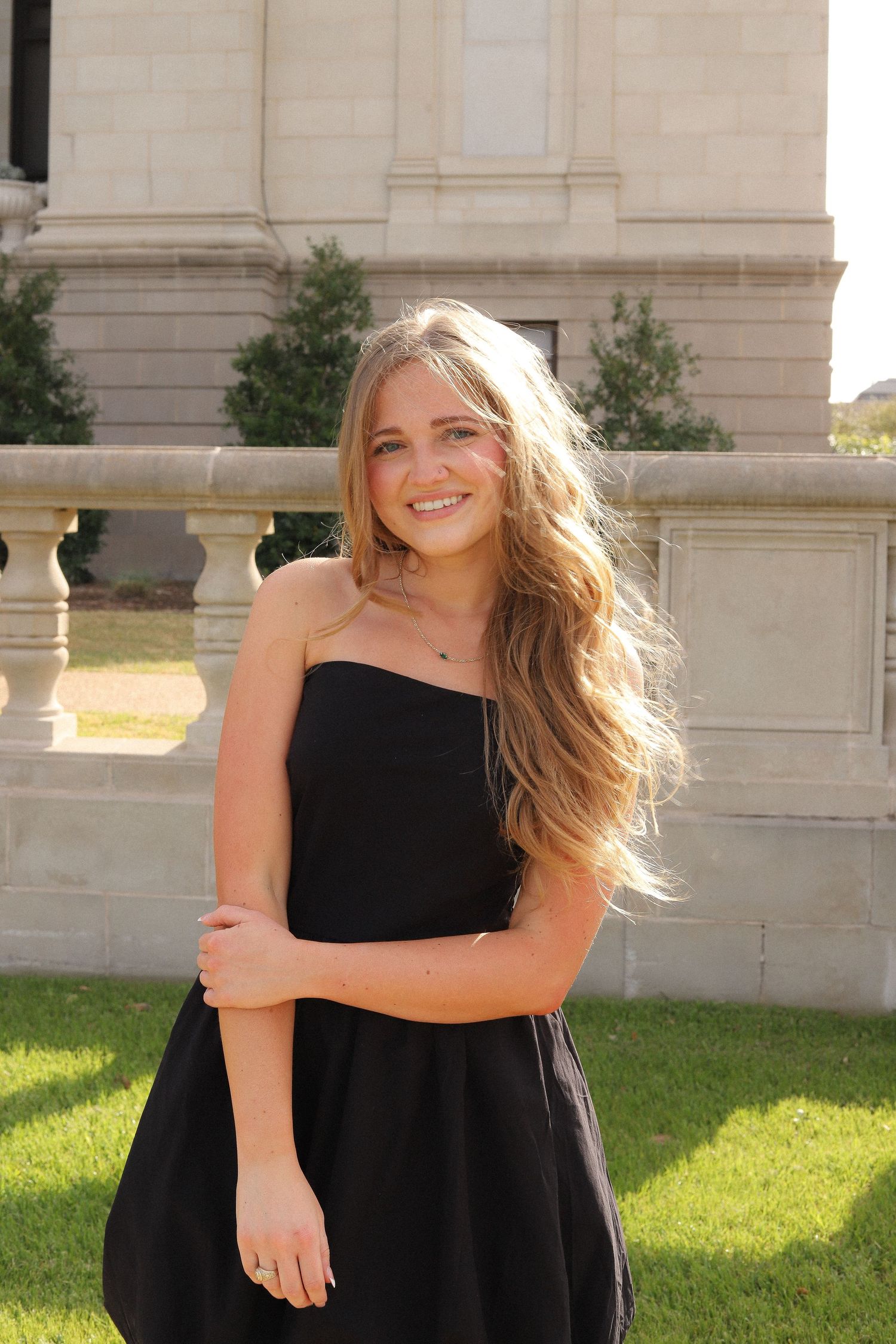 A person in a black strapless dress poses outdoors near a stone building on a sunny day.