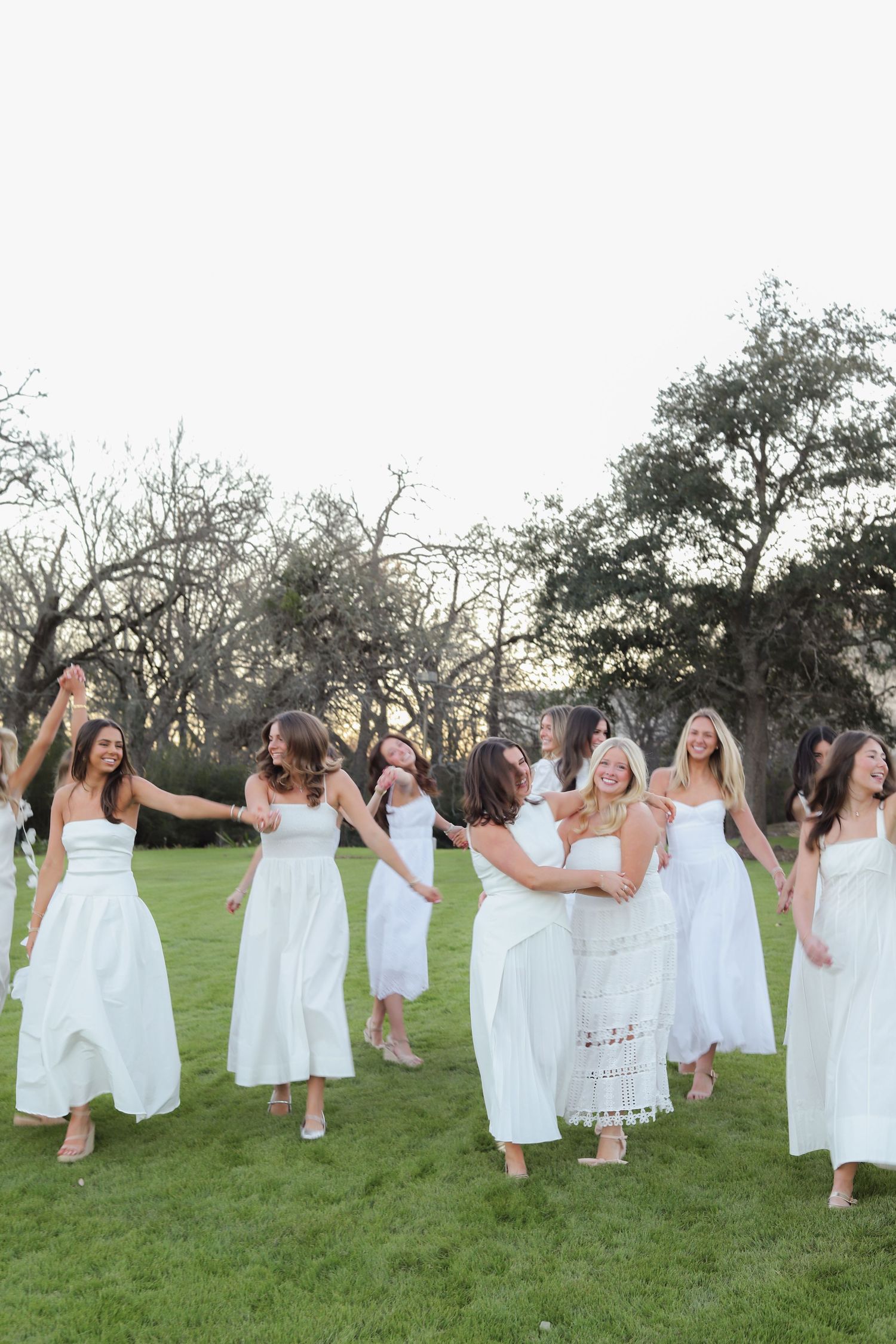 A joyful group in white dresses dance and celebrate together on an outdoor lawn.