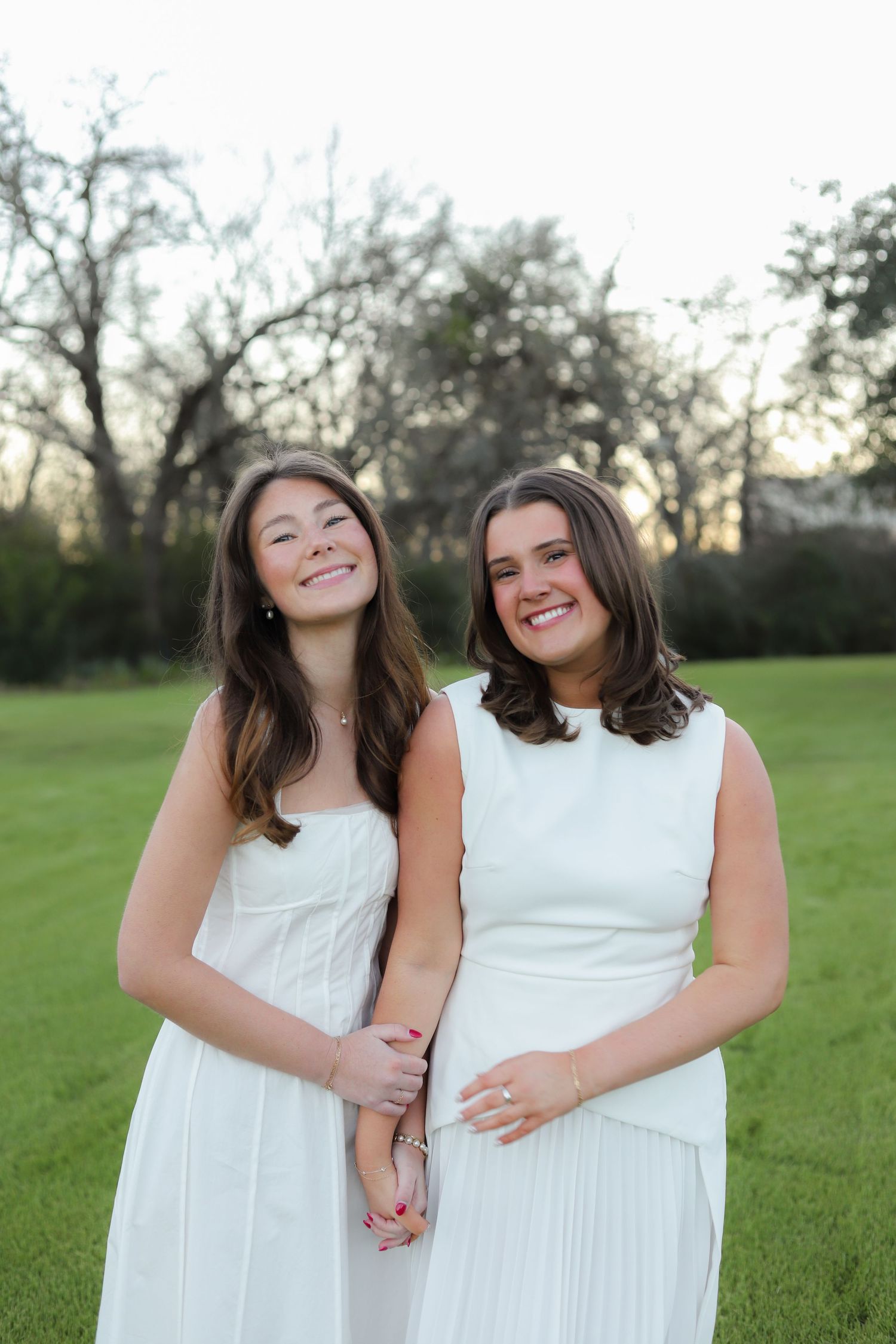 Two women in white dresses share candid moment while laughing in grassy field.