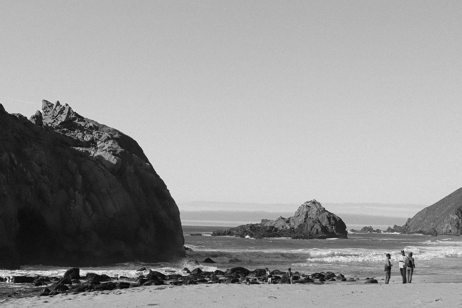 Black and white coastal scene with rock formations and two people walking on a remote beach.
