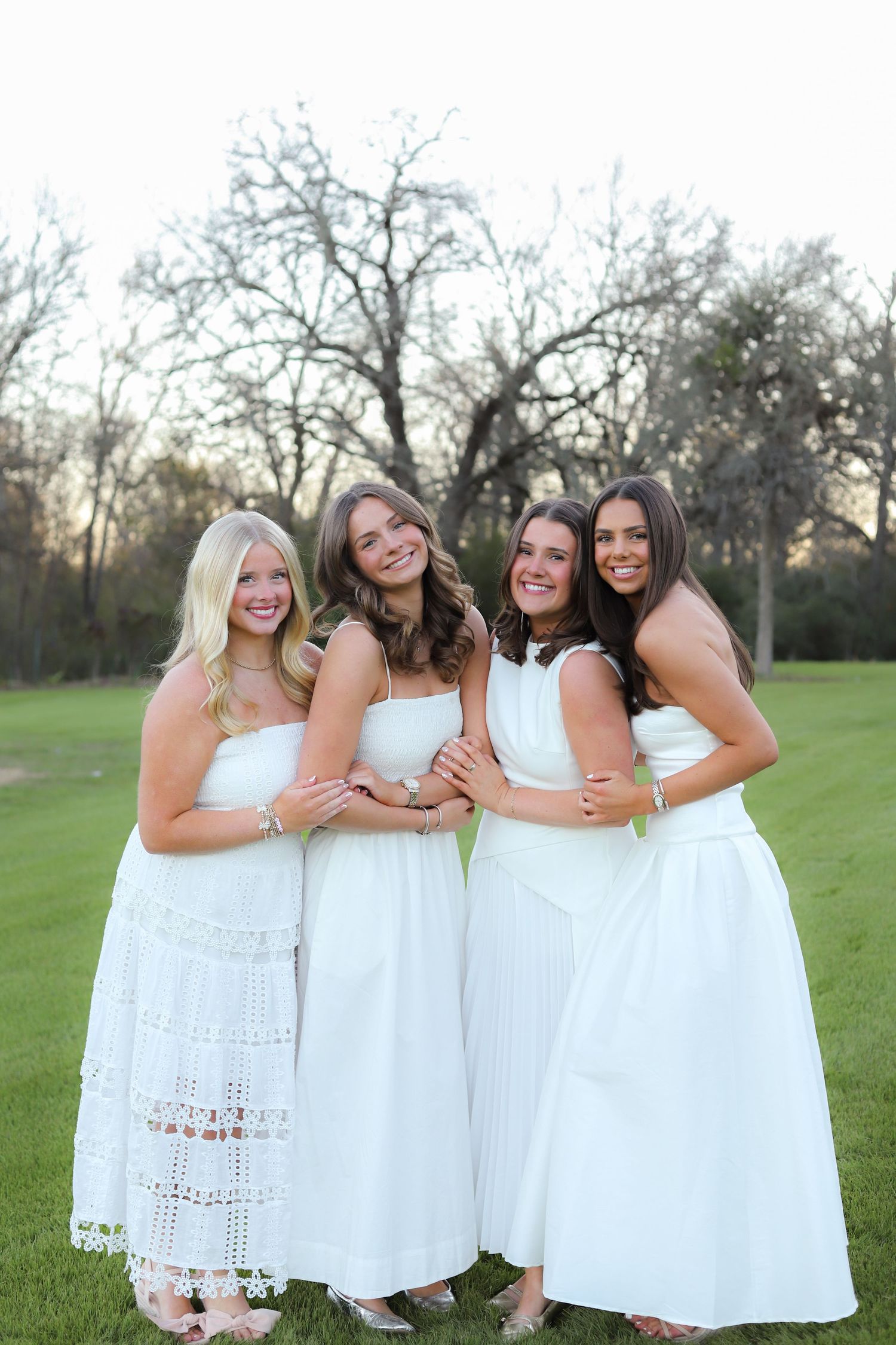 Friends in coordinated white dresses embrace while standing on green grass during a photo session.