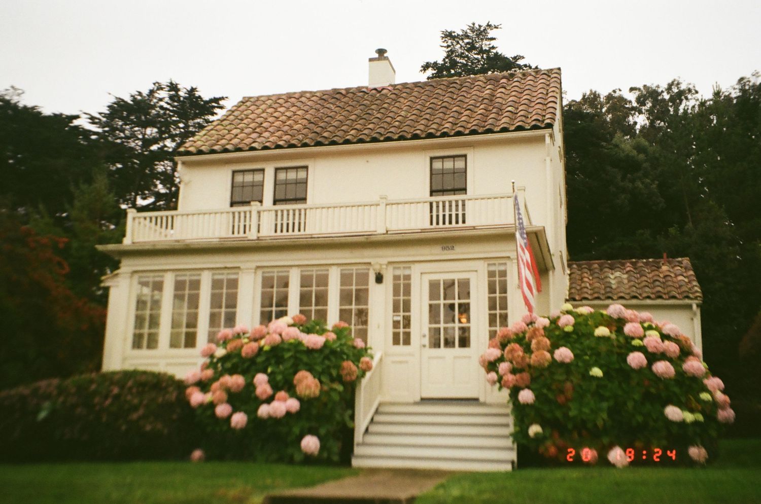 White colonial style house with peach colored roses blooming along the foundation in a lush garden setting.