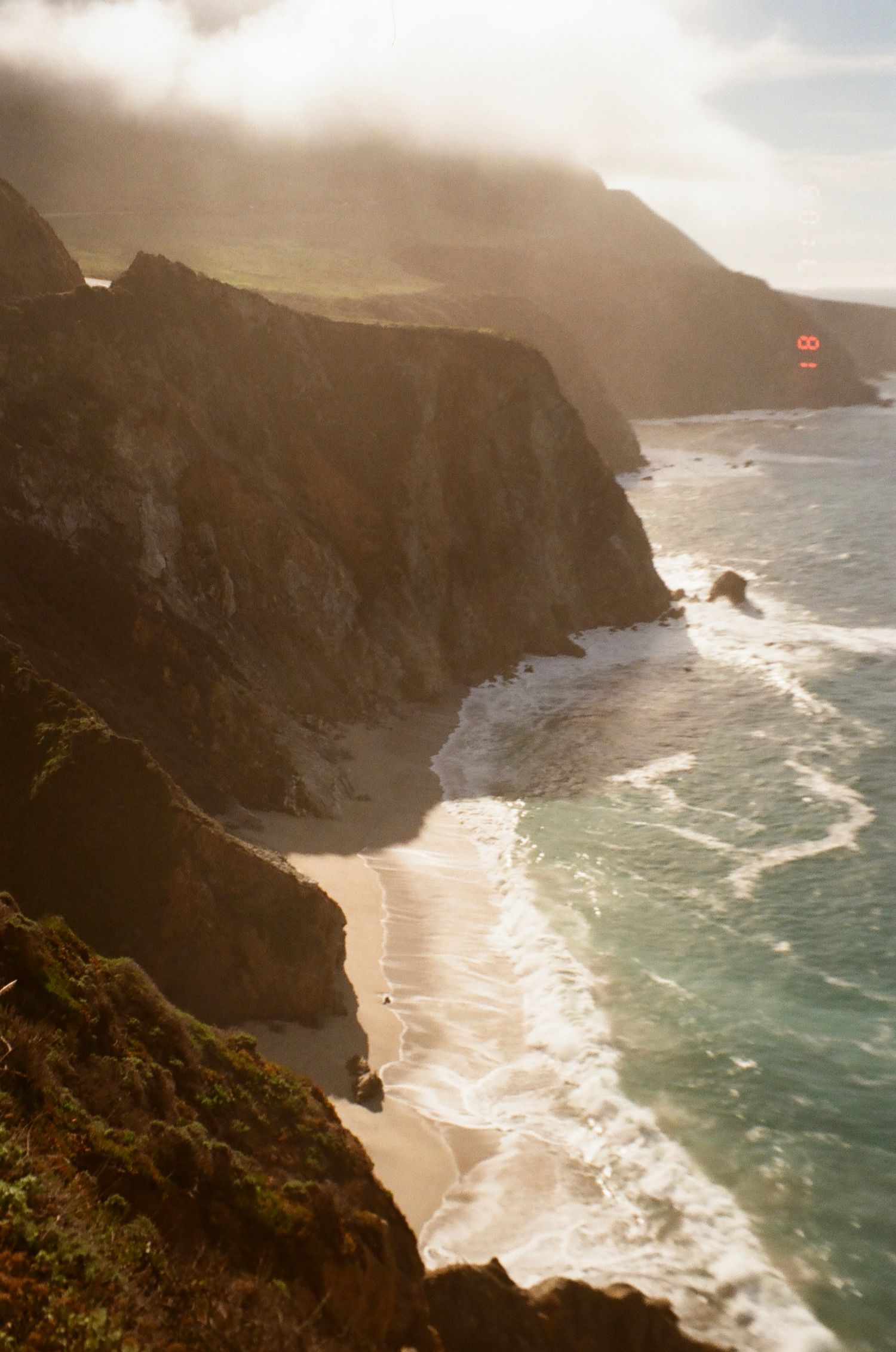 Dramatic coastal cliffs meet the ocean at sunset, with waves crashing against a hidden beach between rocky formations.