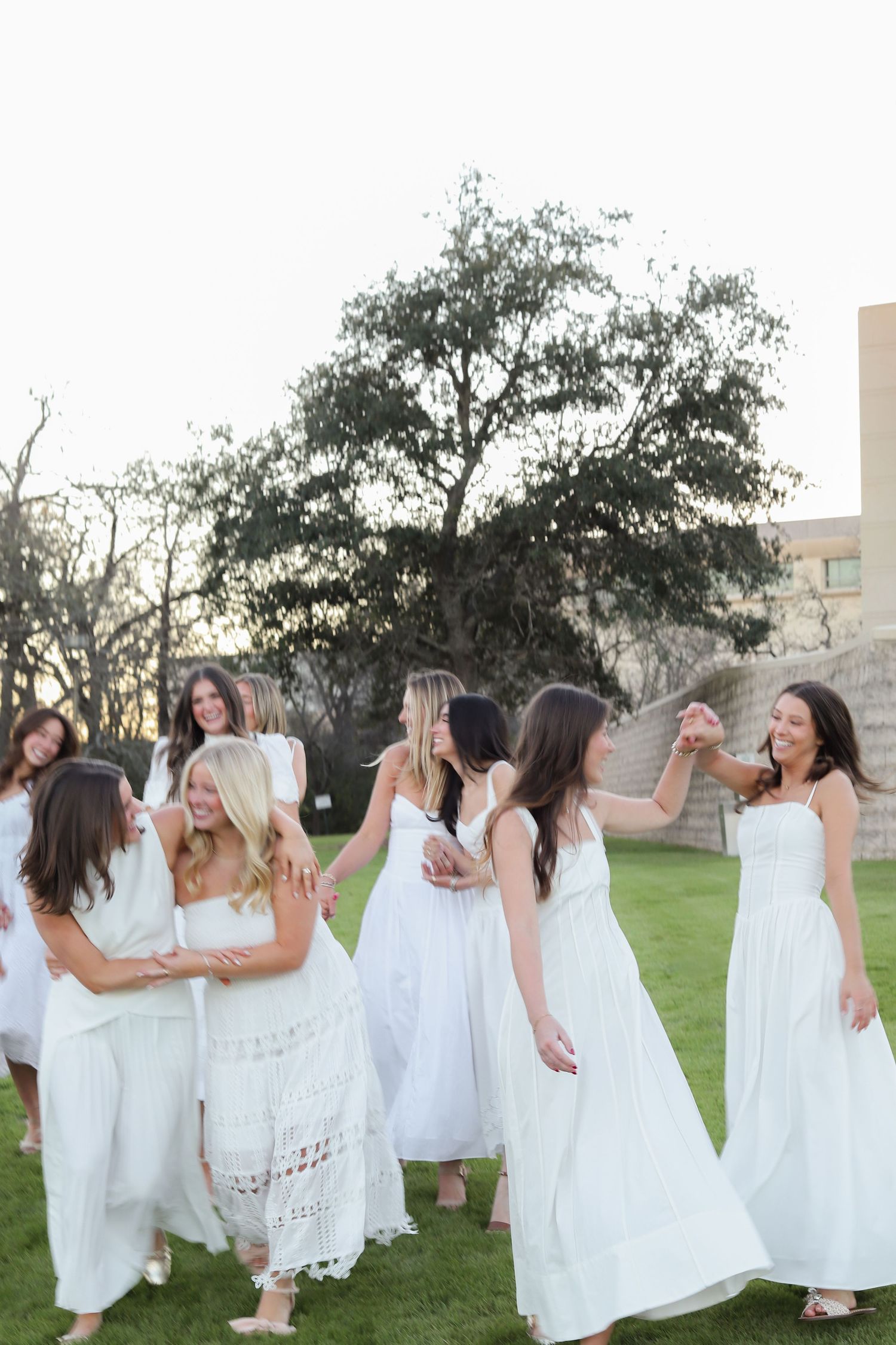 Group of women in white dresses dance and celebrate on grass at dusk.