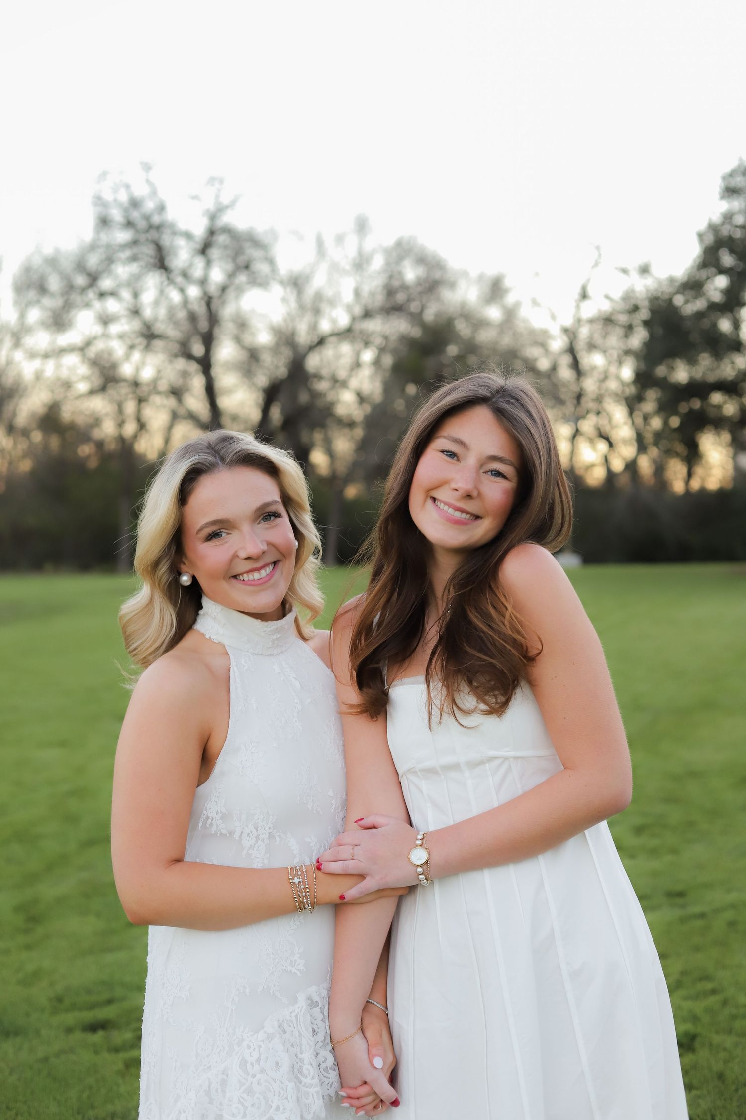 Two people in white lace dresses embrace and smile while standing in a grassy field at sunset.