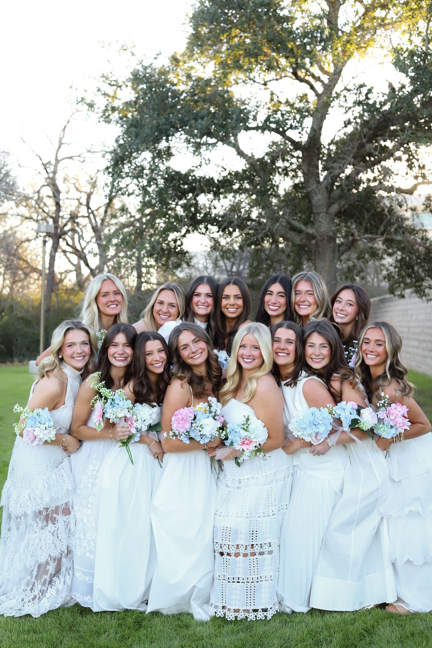 Bridesmaids in white dresses holding pastel bouquets pose together for formal photos in a park setting.