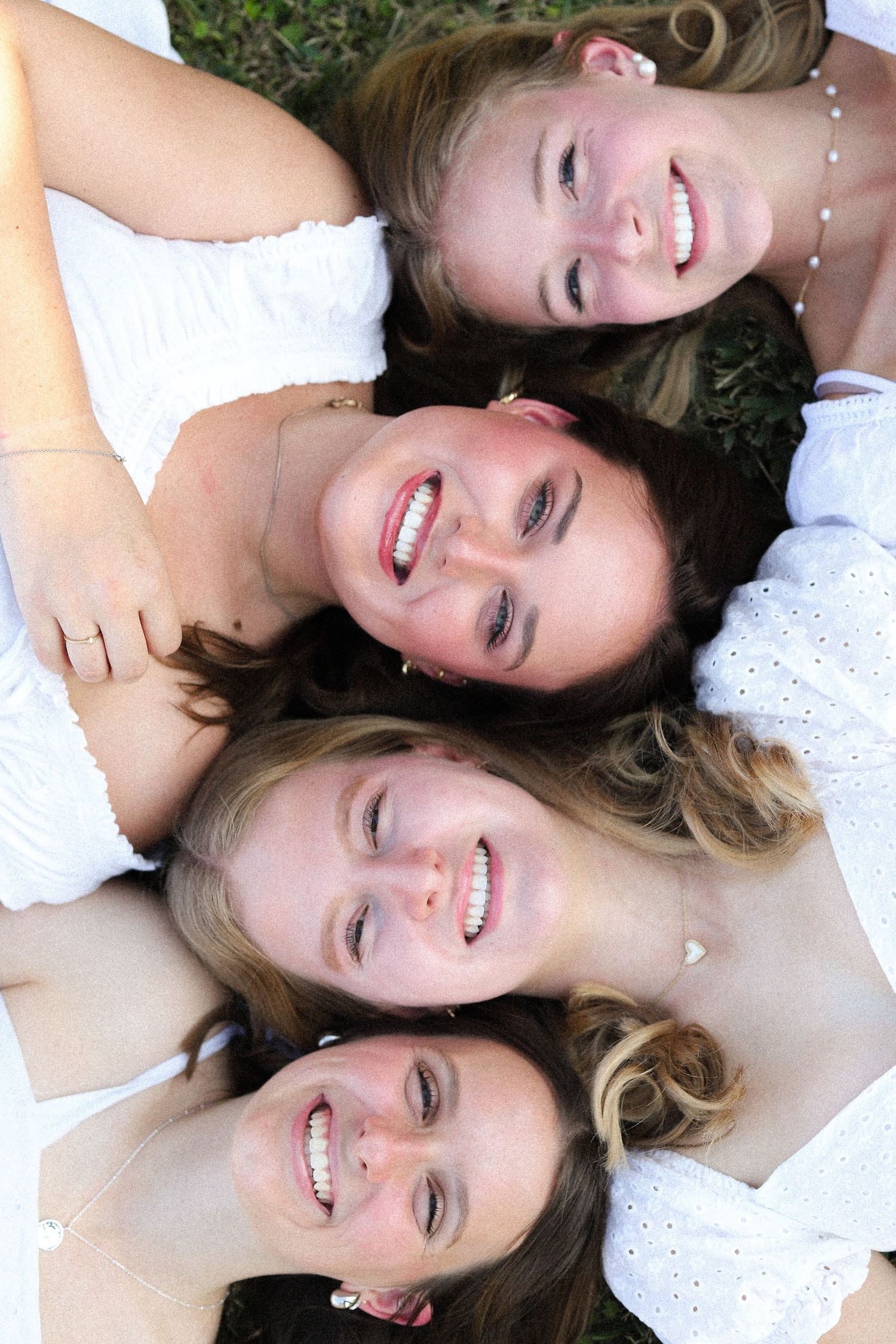 Four friends lying on grass wearing white tops smiling at camera in circle formation.