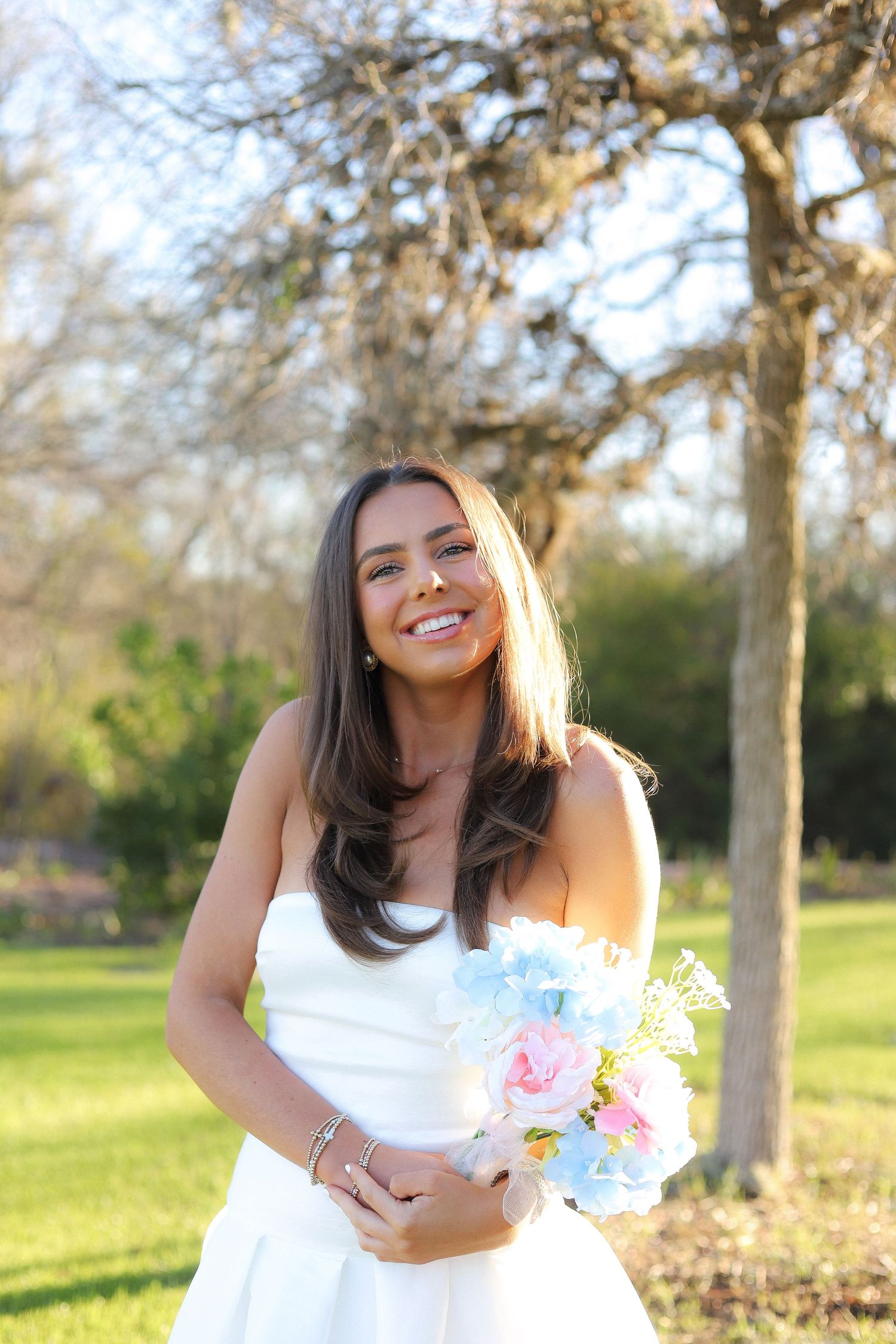 A person in a white dress poses joyfully in a sunny park setting with trees in the background.