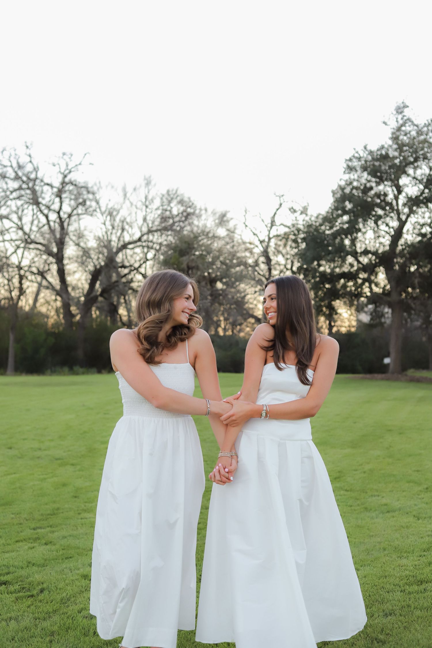 Two friends in flowing white dresses embrace while laughing together in an outdoor setting.