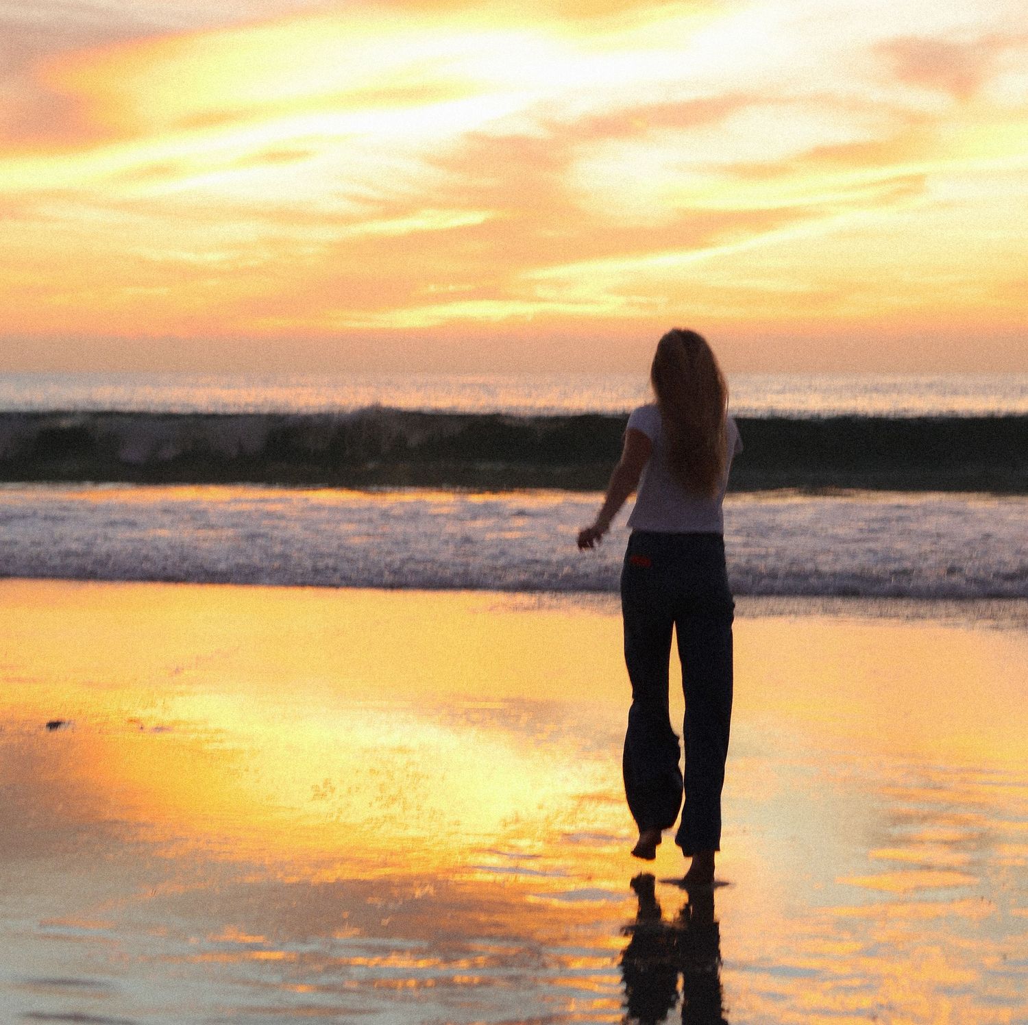 Silhouette of a person standing on a reflective beach at sunset with golden clouds and crashing waves.