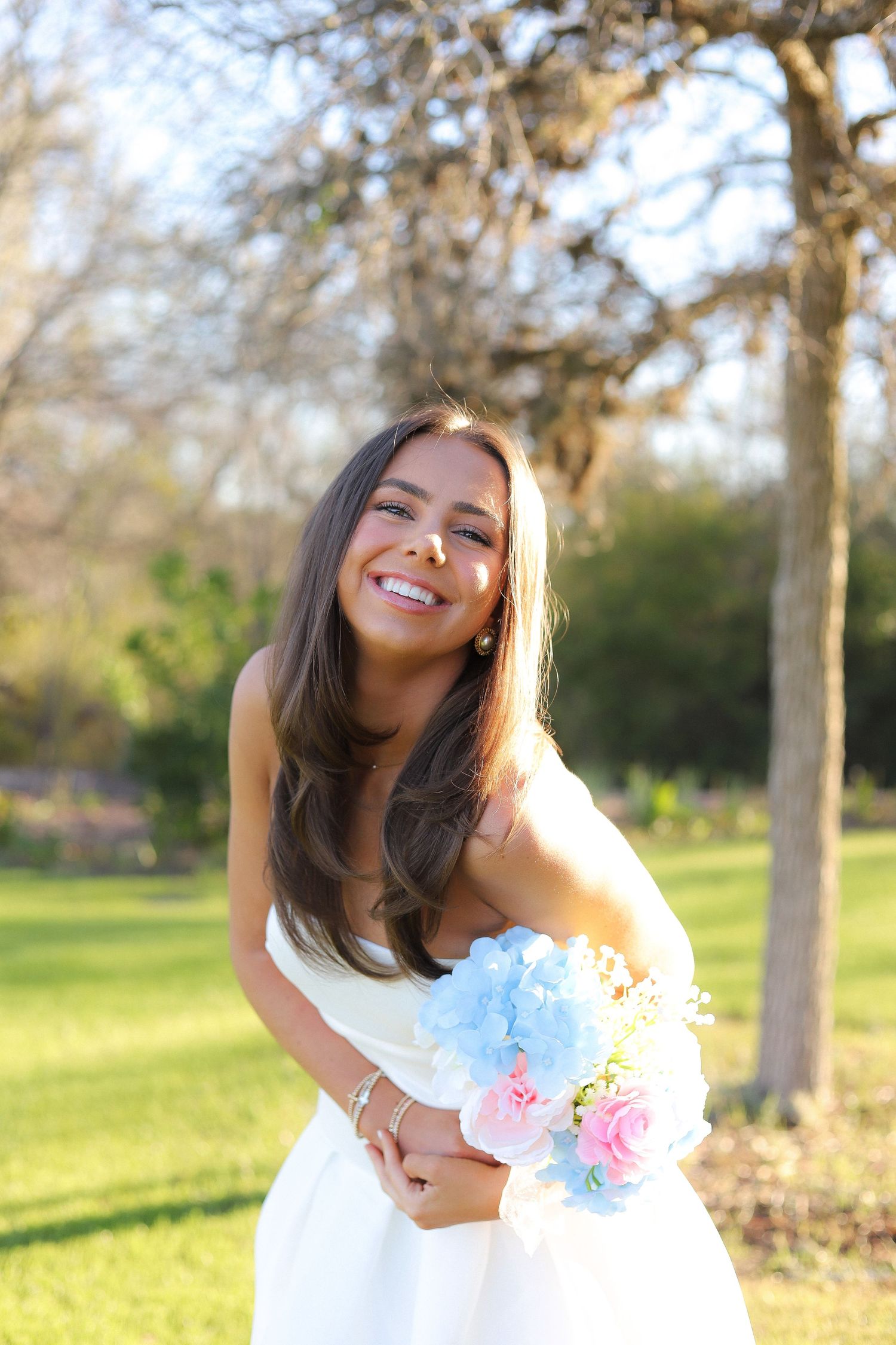 A joyful person in a white dress holds a bouquet of pink and blue flowers in a sunny outdoor setting.