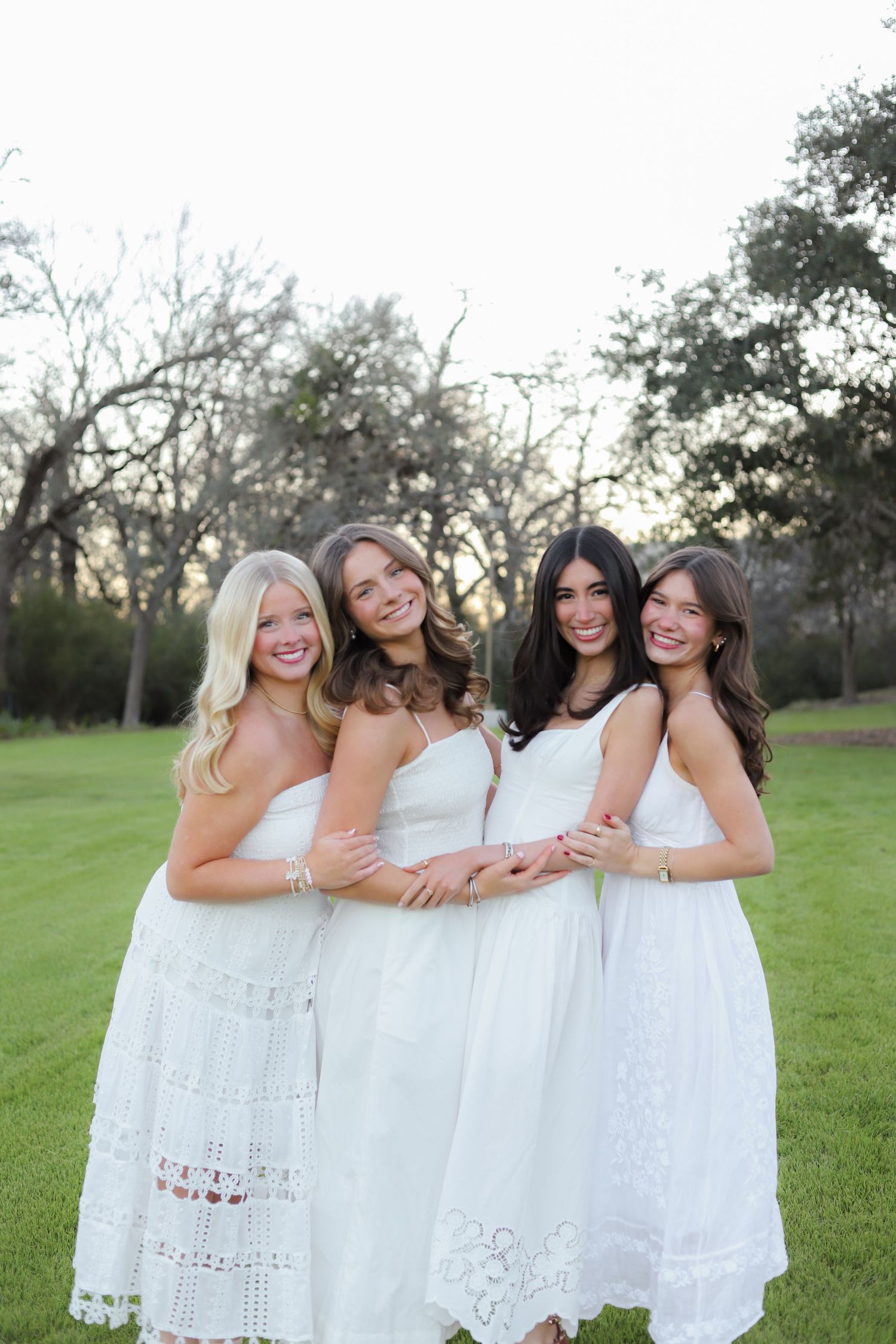 Friends in matching white dresses embrace while standing on grass at sunset.