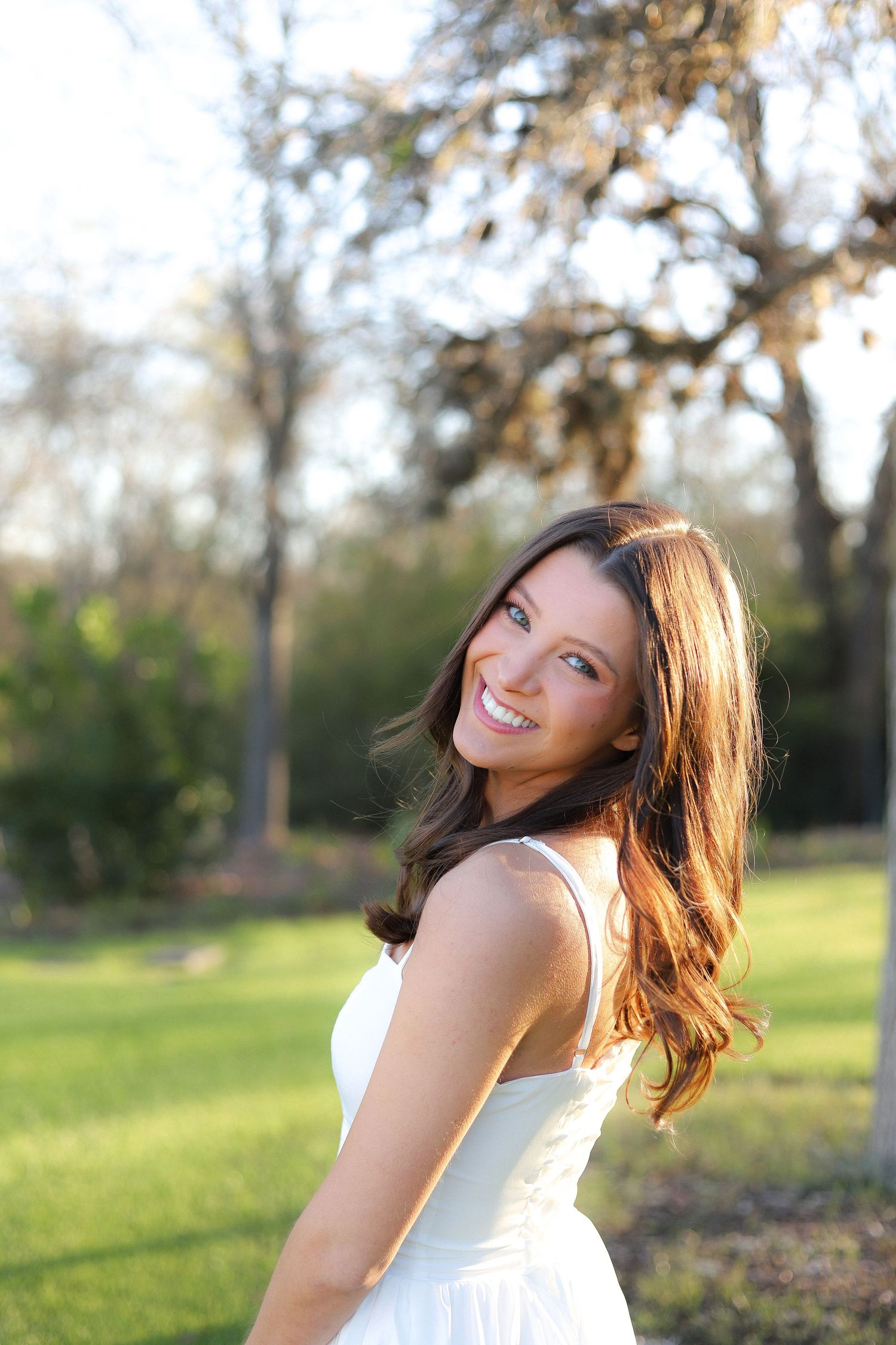 A smiling figure in a white dress poses outdoors in a grassy area with trees during golden hour lighting.