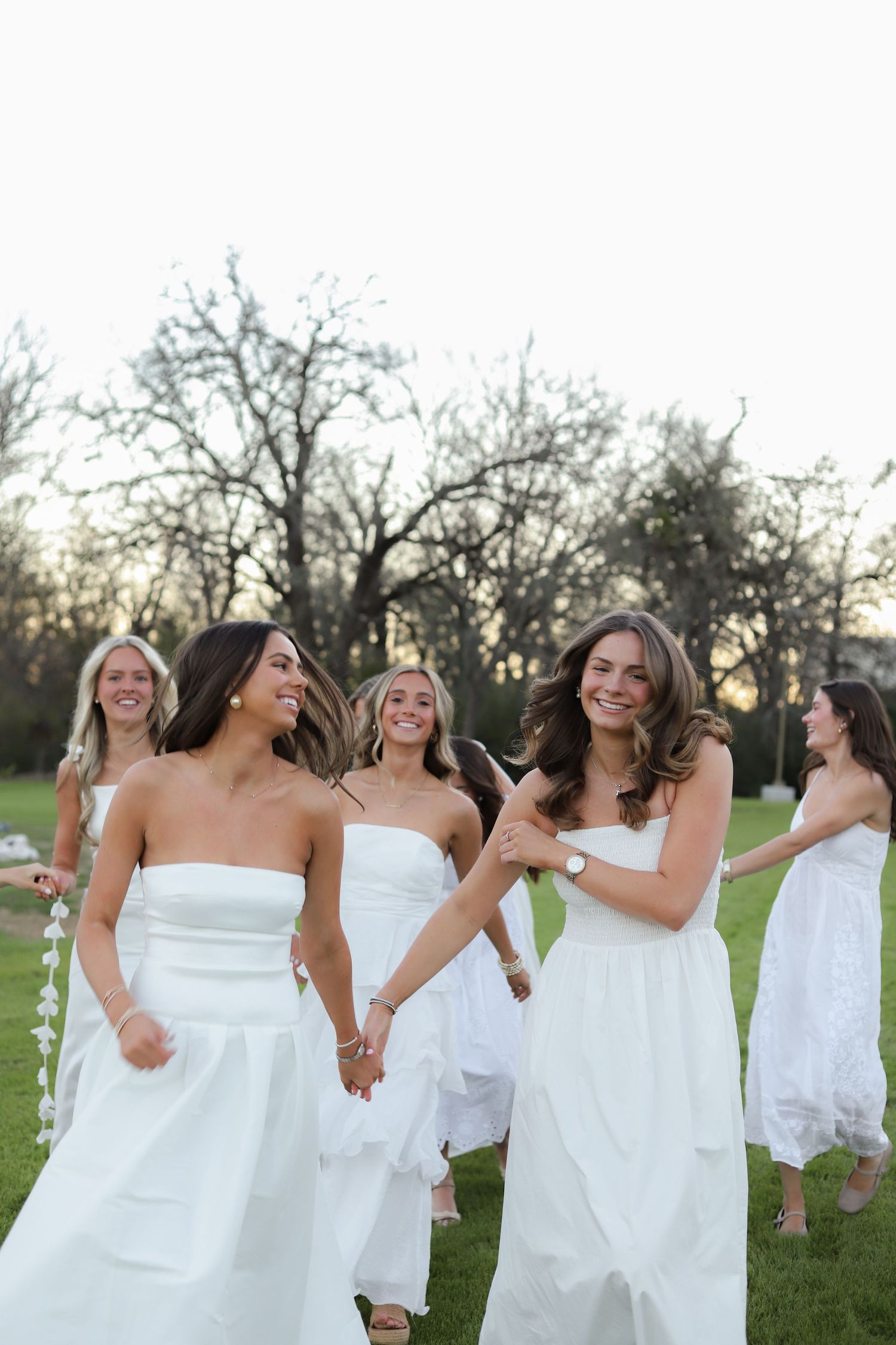 Women in flowing white dresses laugh together while holding hands on a lawn at sunset.
