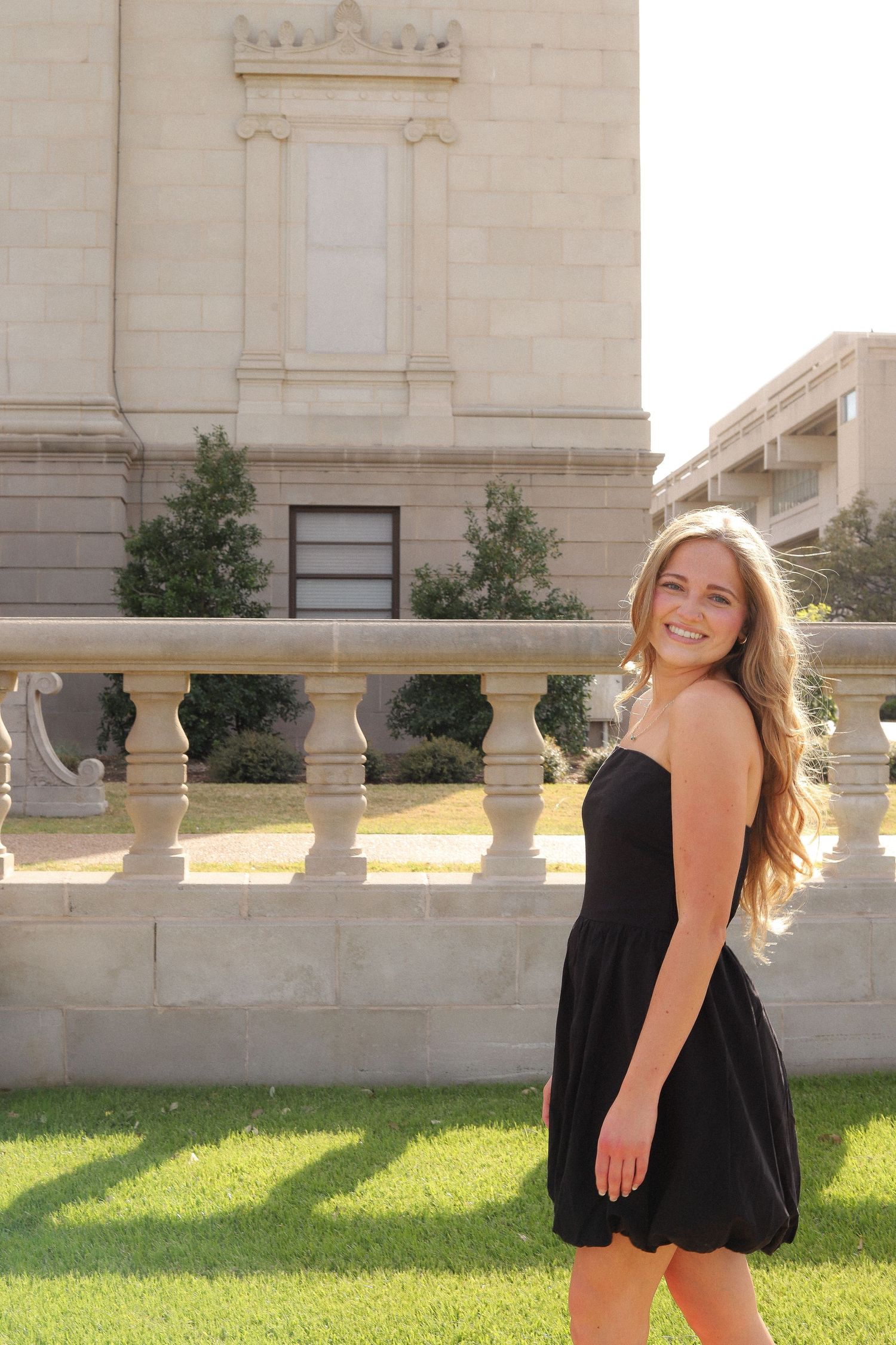 Figure in black dress poses joyfully on stone terrace with classical architecture in background.
