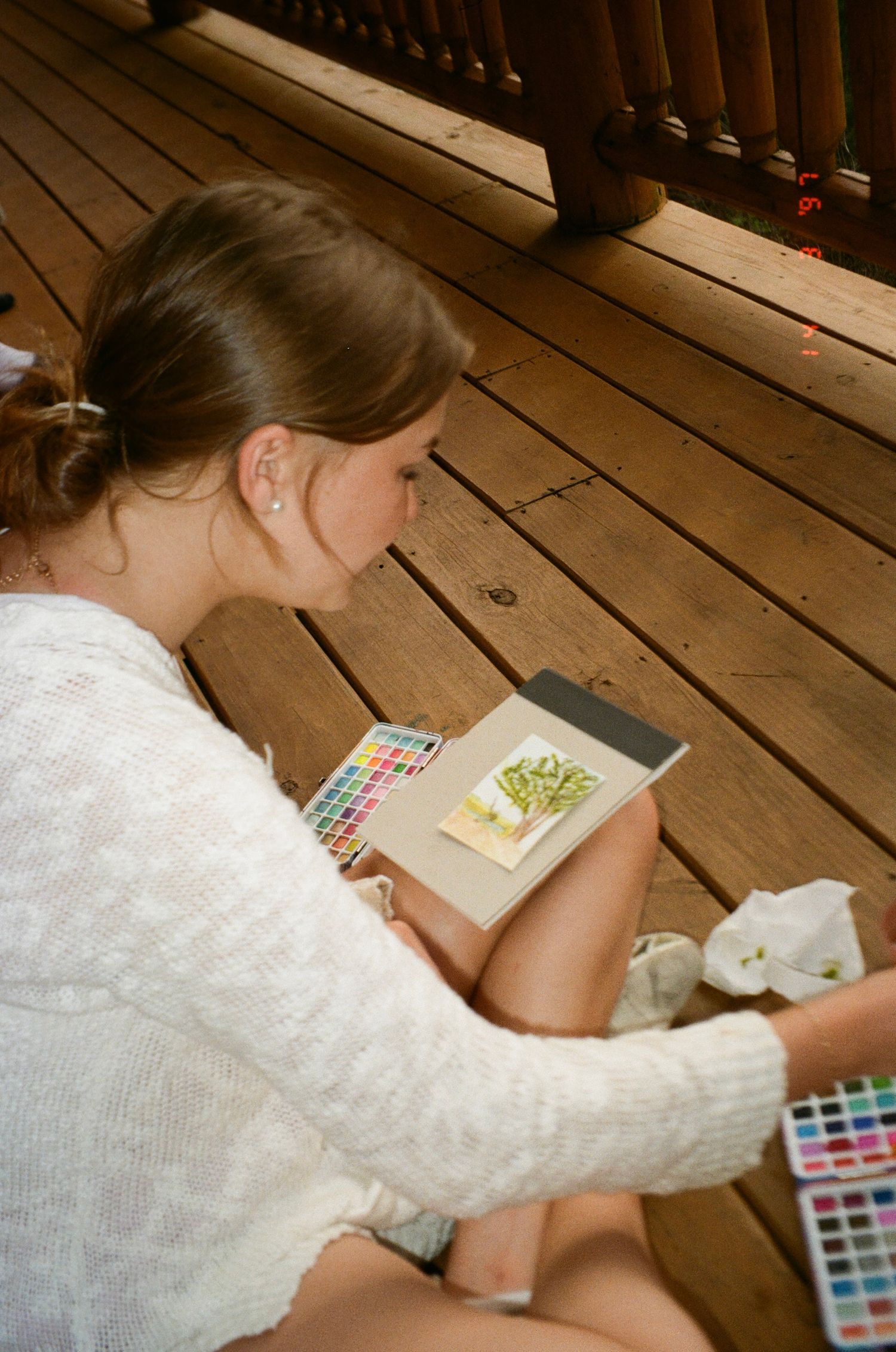 Artist working with watercolor palette and sketchbook on wooden deck creates colorful artwork in natural lighting.