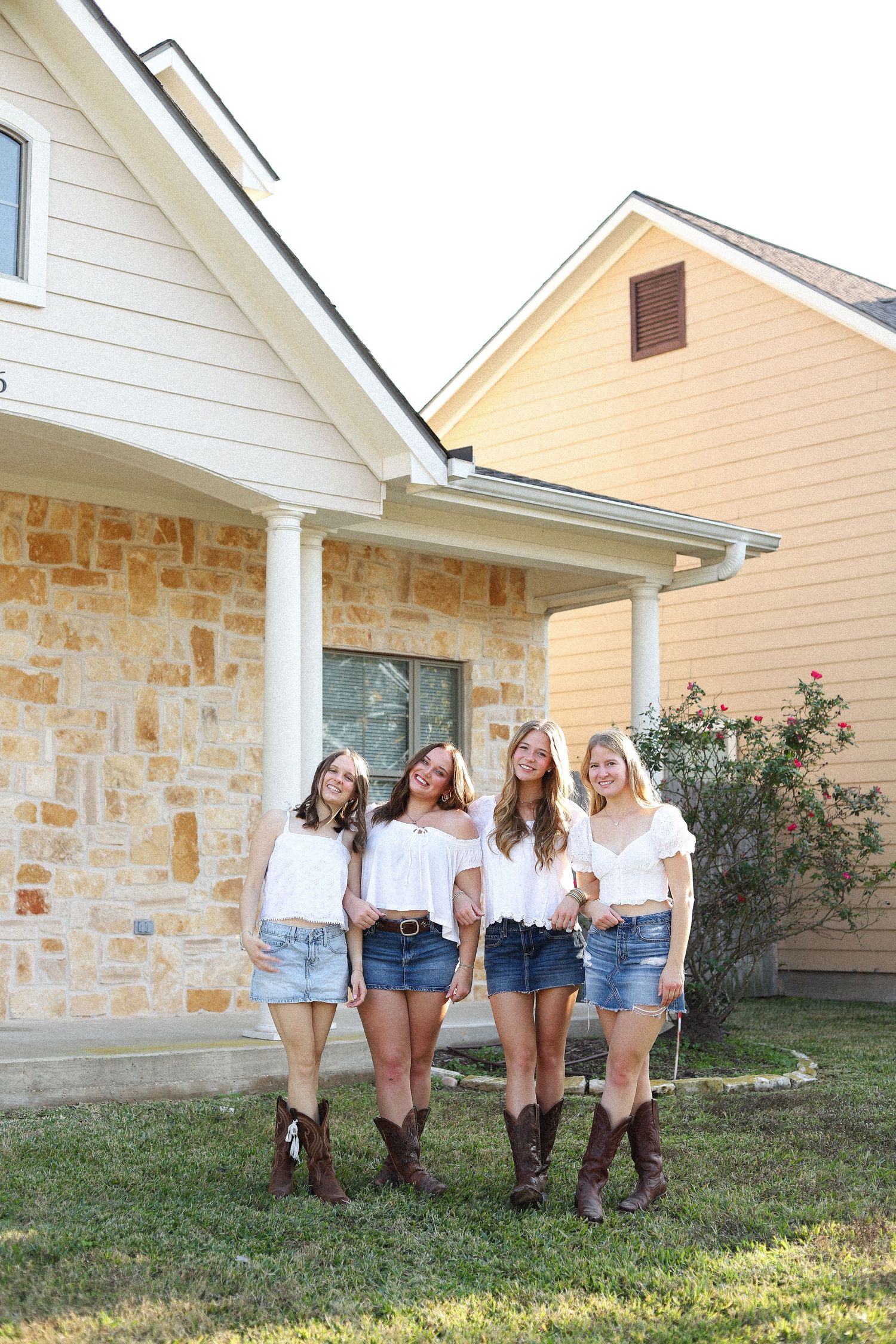 A group poses together wearing matching white tops, denim shorts and brown boots against a stone house exterior.