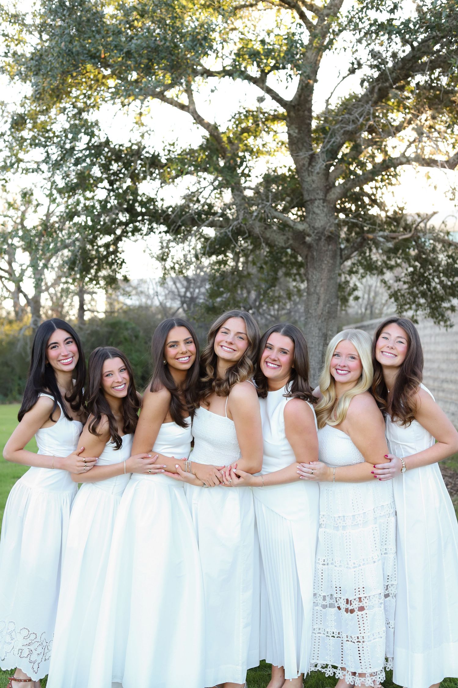 A group of friends in matching white dresses pose together outdoors for a cheerful group photo.