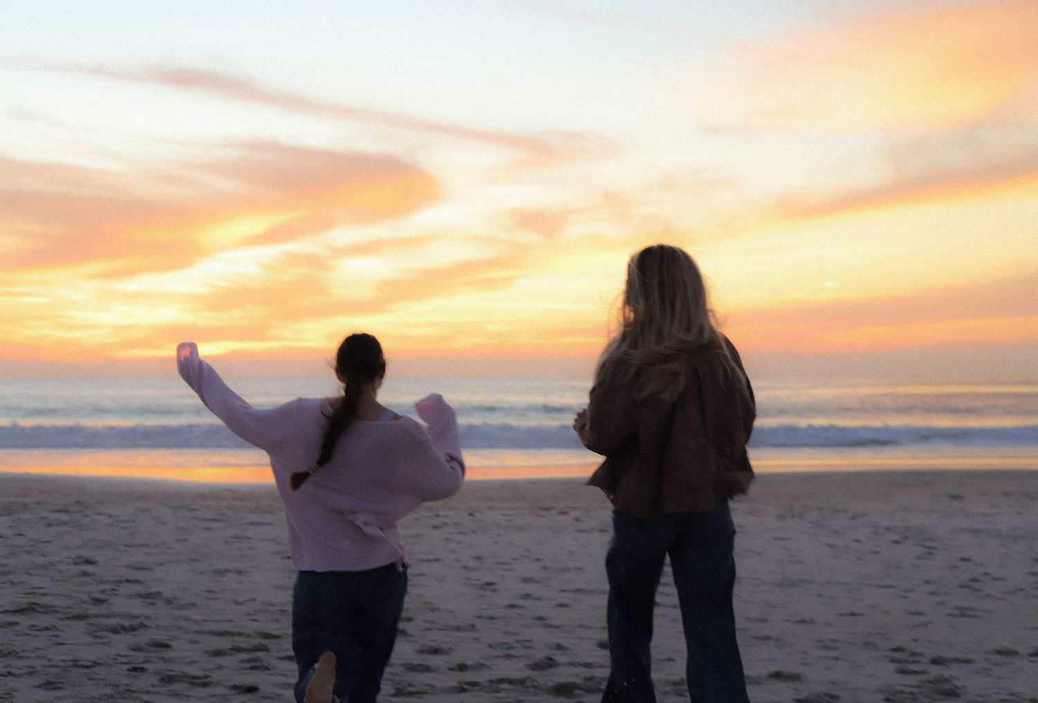 Two people stand with outstretched arms watching a vibrant orange sunset over the beach.