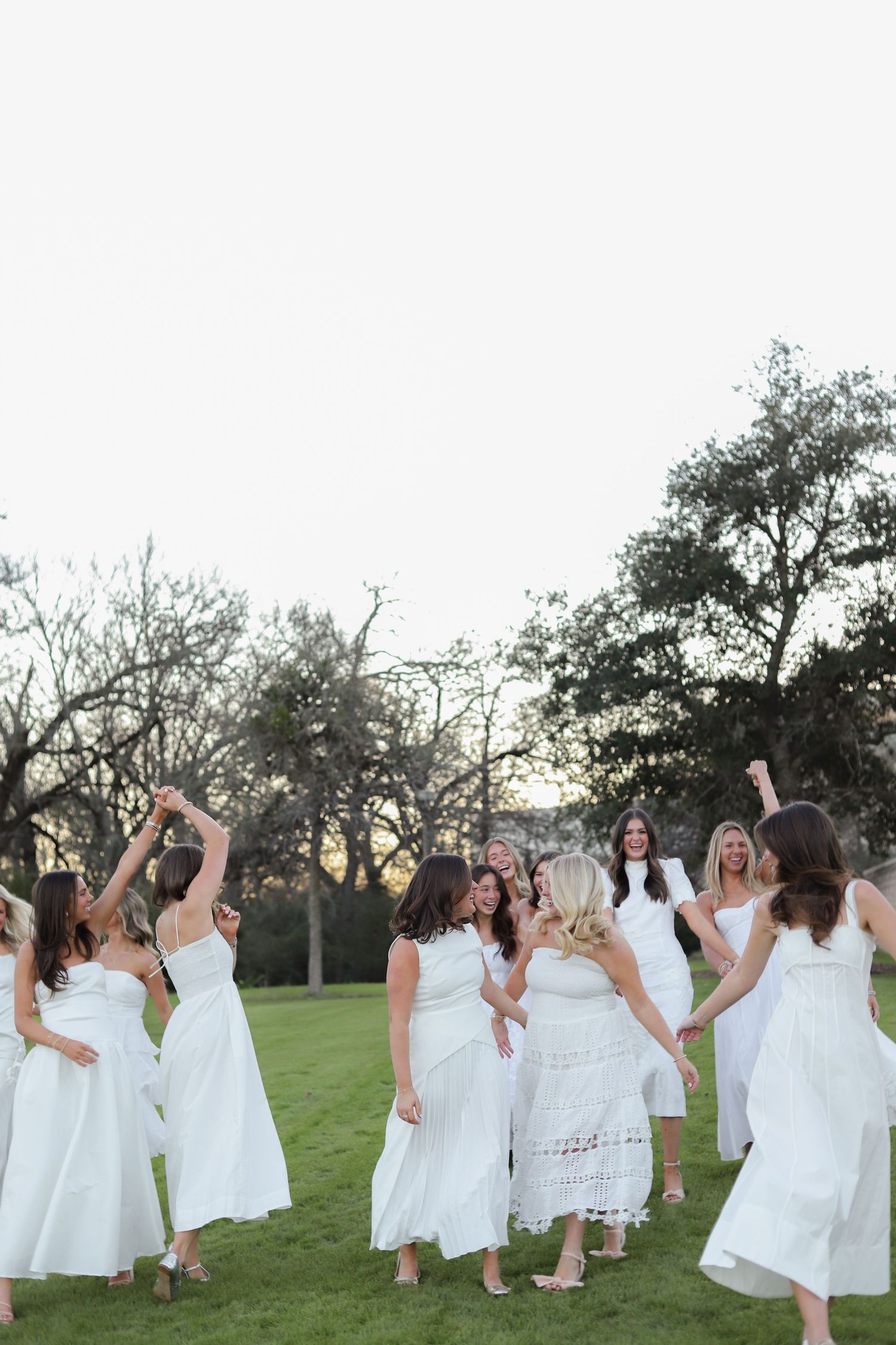 Women in white dresses jump and celebrate together on a green lawn during golden hour.