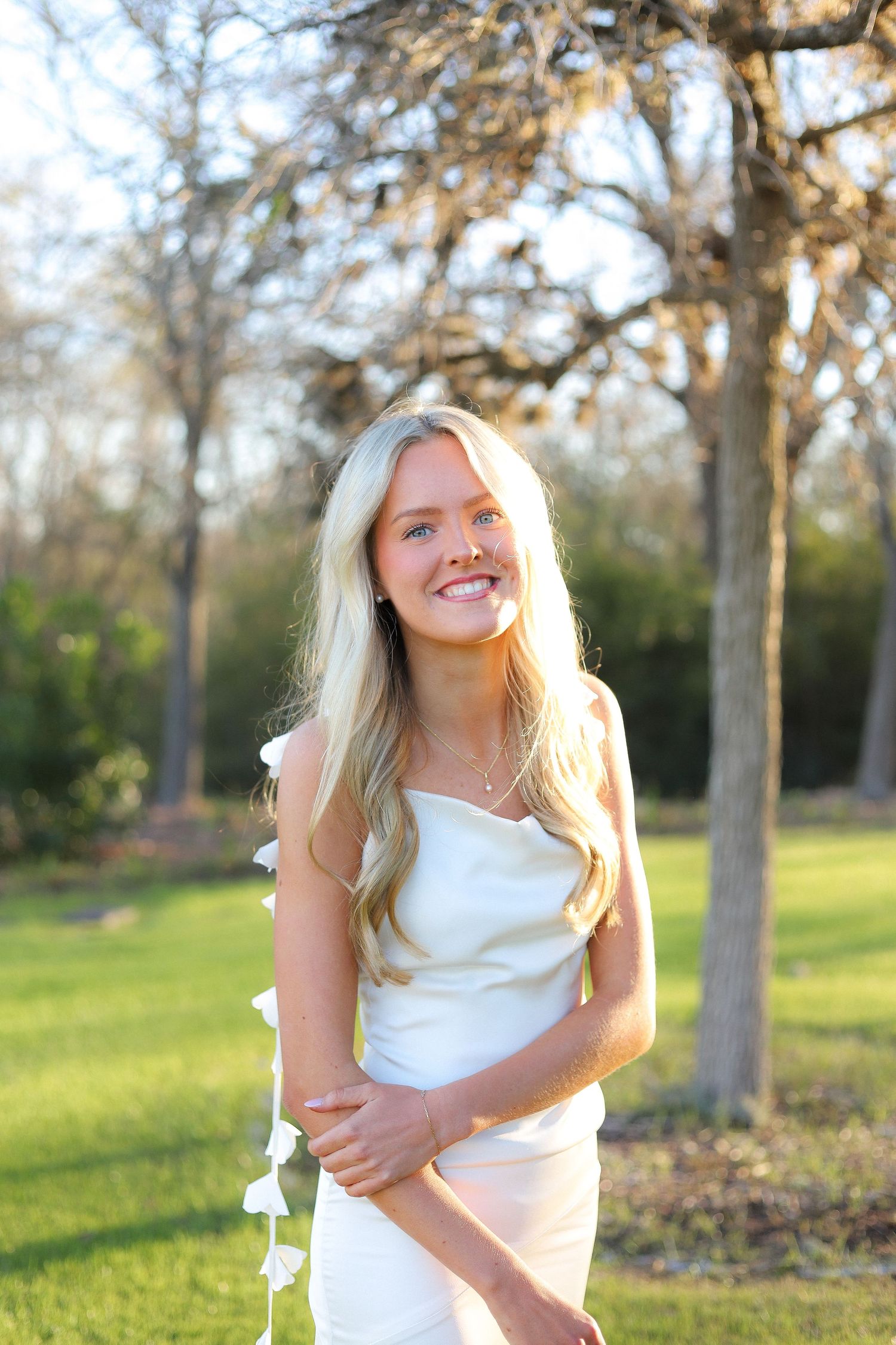 A person in a white dress poses playfully in a sunny outdoor setting with trees in the background.