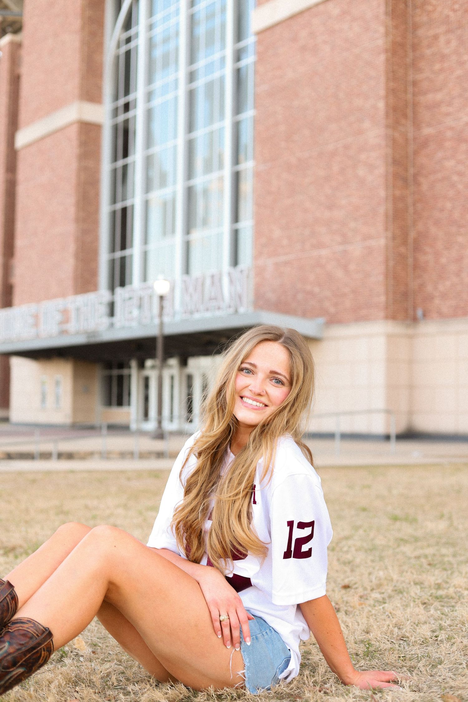Student in white jersey number 12 and denim shorts posing on grass near brick building.