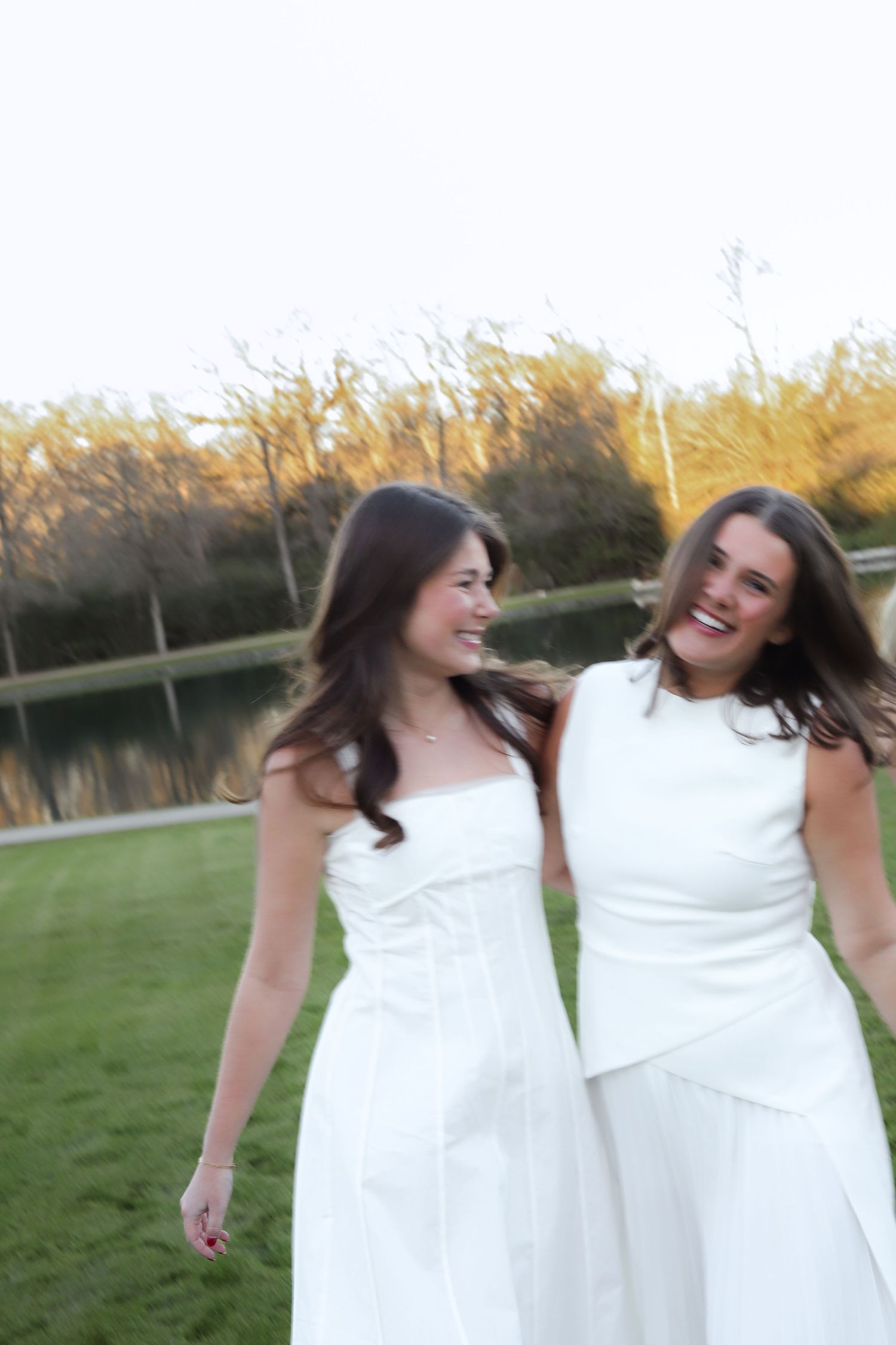 Two women in white dresses laugh together while standing on grass near a pond at sunset.